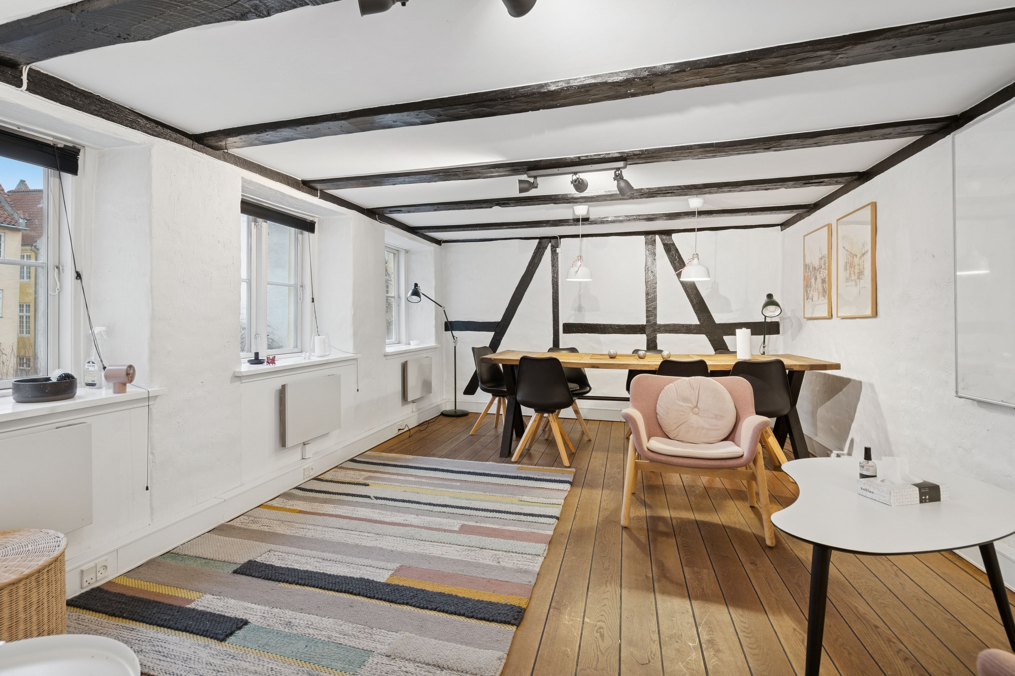 Kontor med bord og stole samt en sofa med pude i et lyse, moderne og funktionelt kontor med loftsbjælker og trægulv på Sankt Peders Stræde 39, 2. sal, København K