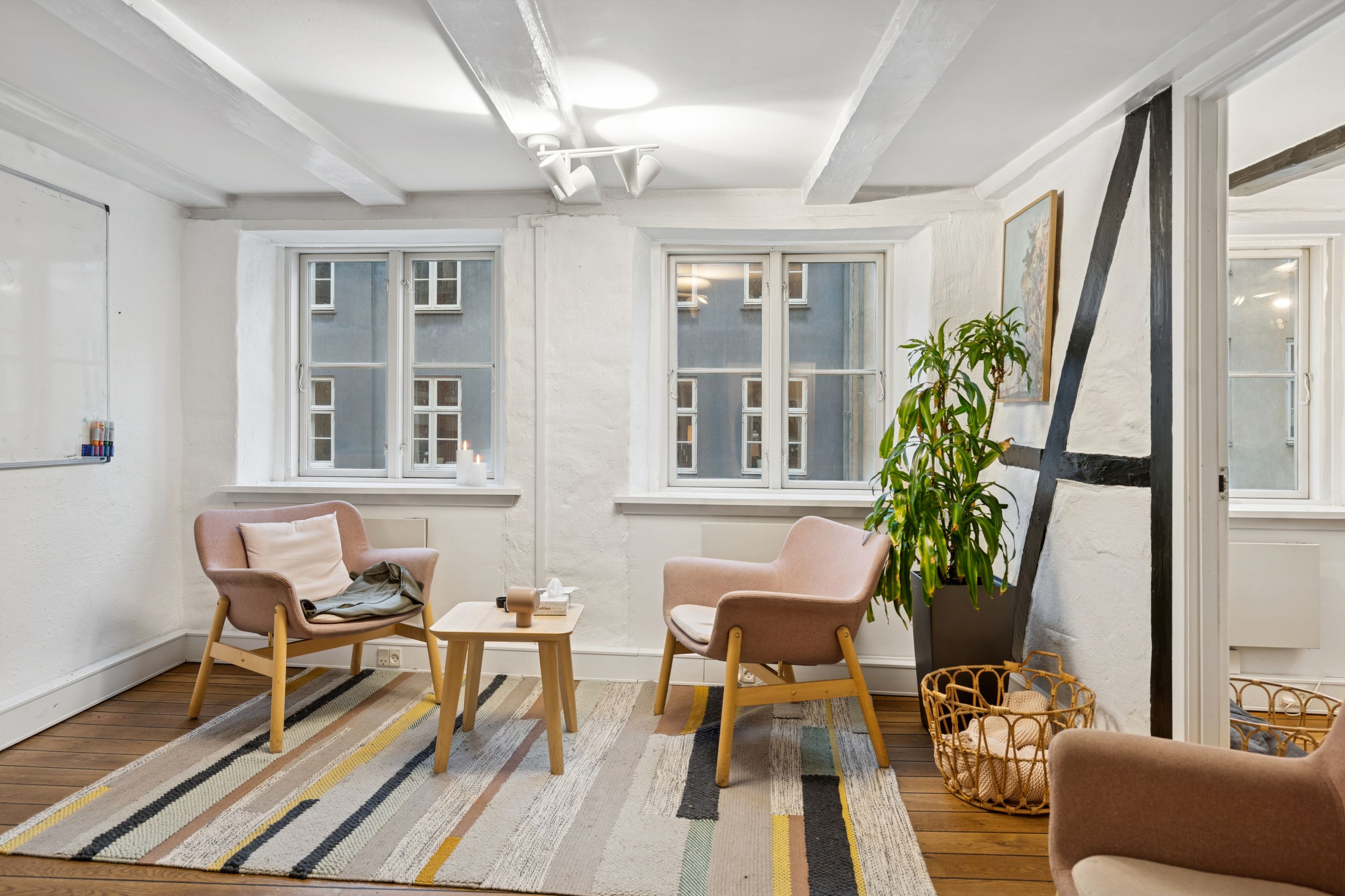 Kontorrum med moderne møbler, hvid væg og vinduer med udsigt til bygninger og lyse himmel. På gulvet ligger et tæppe og der er en plante i et kurv.