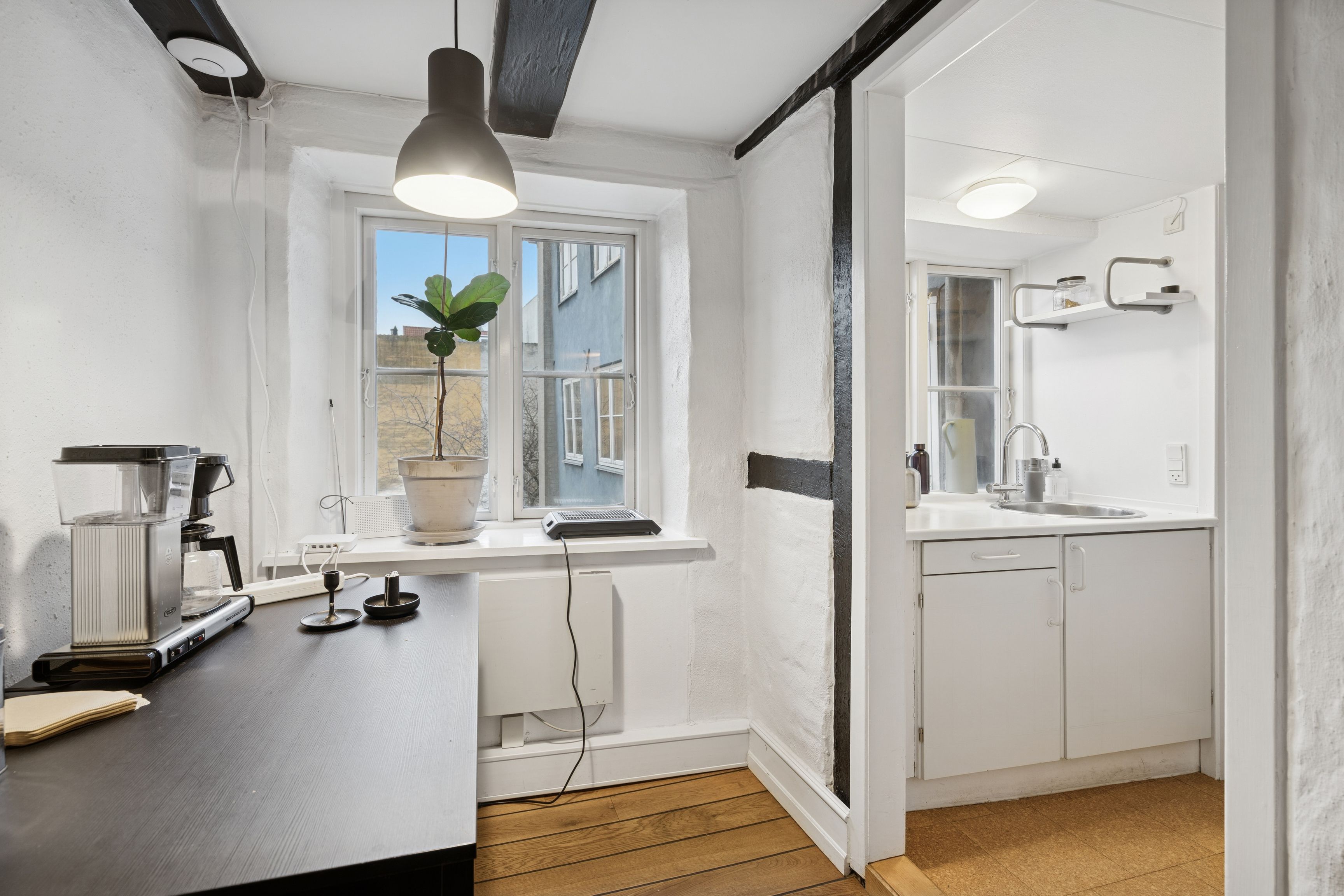 Funktionelt kontor med lyse hjørner og udsigt til byen. Køkken med moderne hvide skabe og en lille kaffemaskine på skrivebordet. En plante i potte og et lysekron.