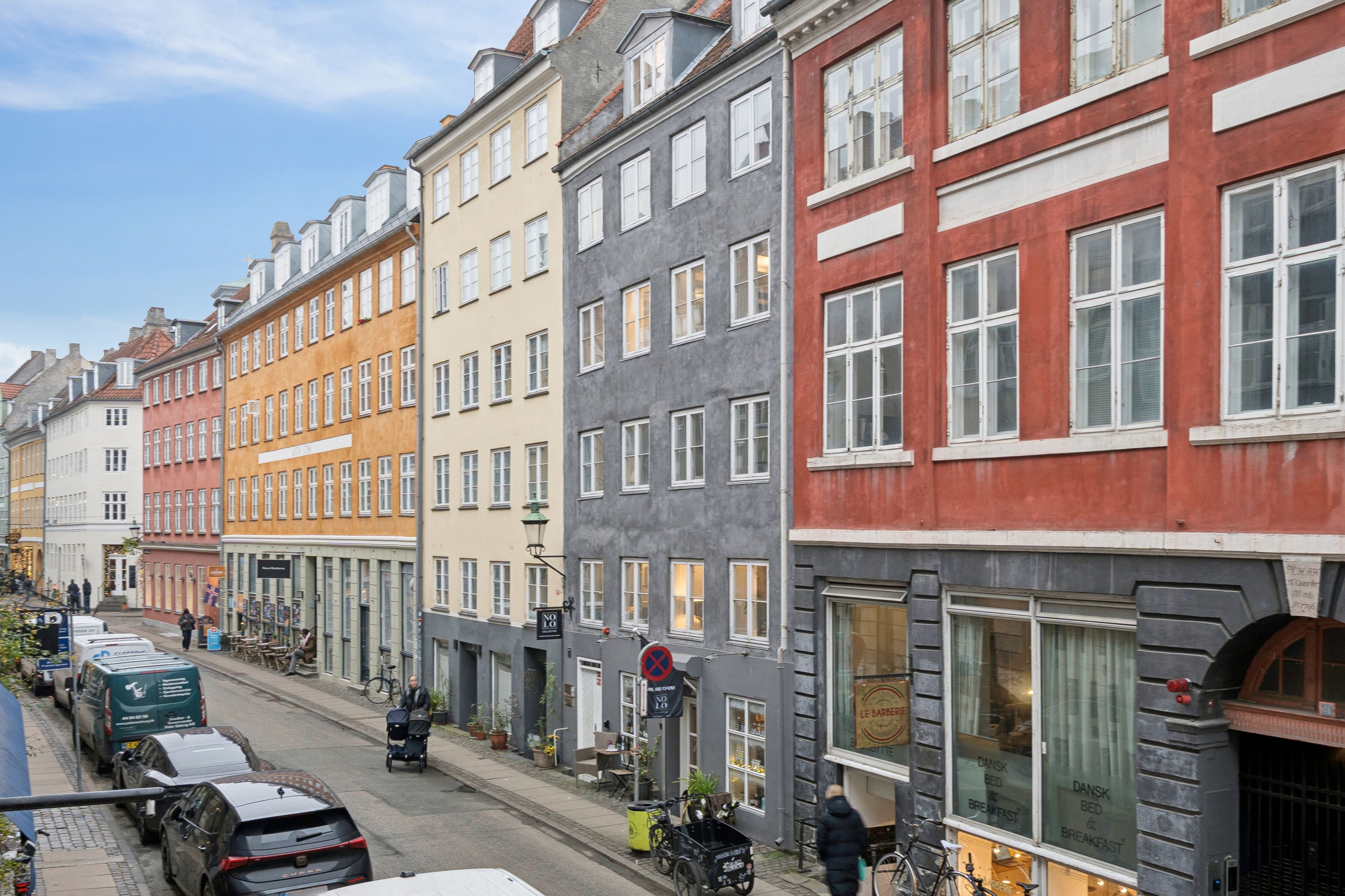 En række farvede bygninger med gadegående butikker og kørende biler på Sankt Peders Stræde i København K. En person kører en barnevogne og en cykel står parkeret på fortauet.