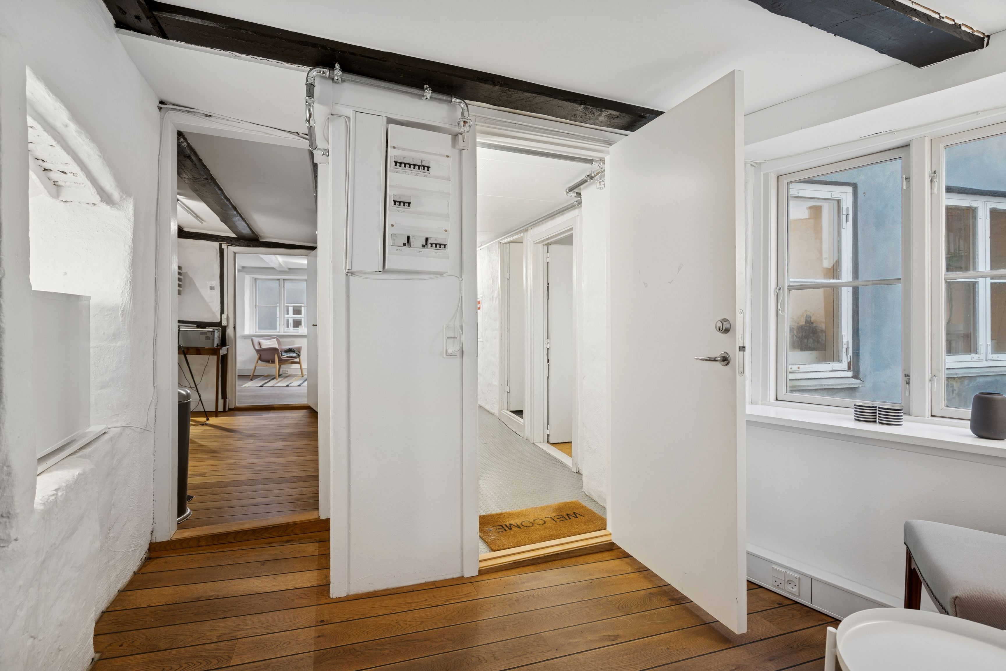 Minimalistisk og velindrettet stue med åbne døre og lyse trægulve, lys og funktionelle indretninger og et vindue med udsigt til byen