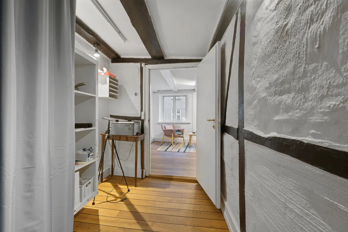 Inde i en velindrettet kontor er der et højt loft med bjælker, en hvid væg og trægulve. Et stativ står med en printer og et skrivebord med en kuffert og en kurv.