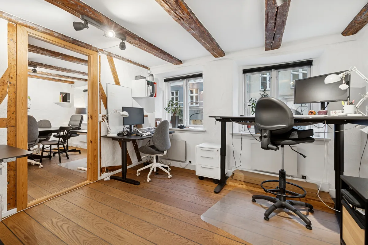 Lyst og velindrettet kontor med højloft og træbjælker, skriveborde og kontorstolar, computere og lamper, og en hvid indretning.