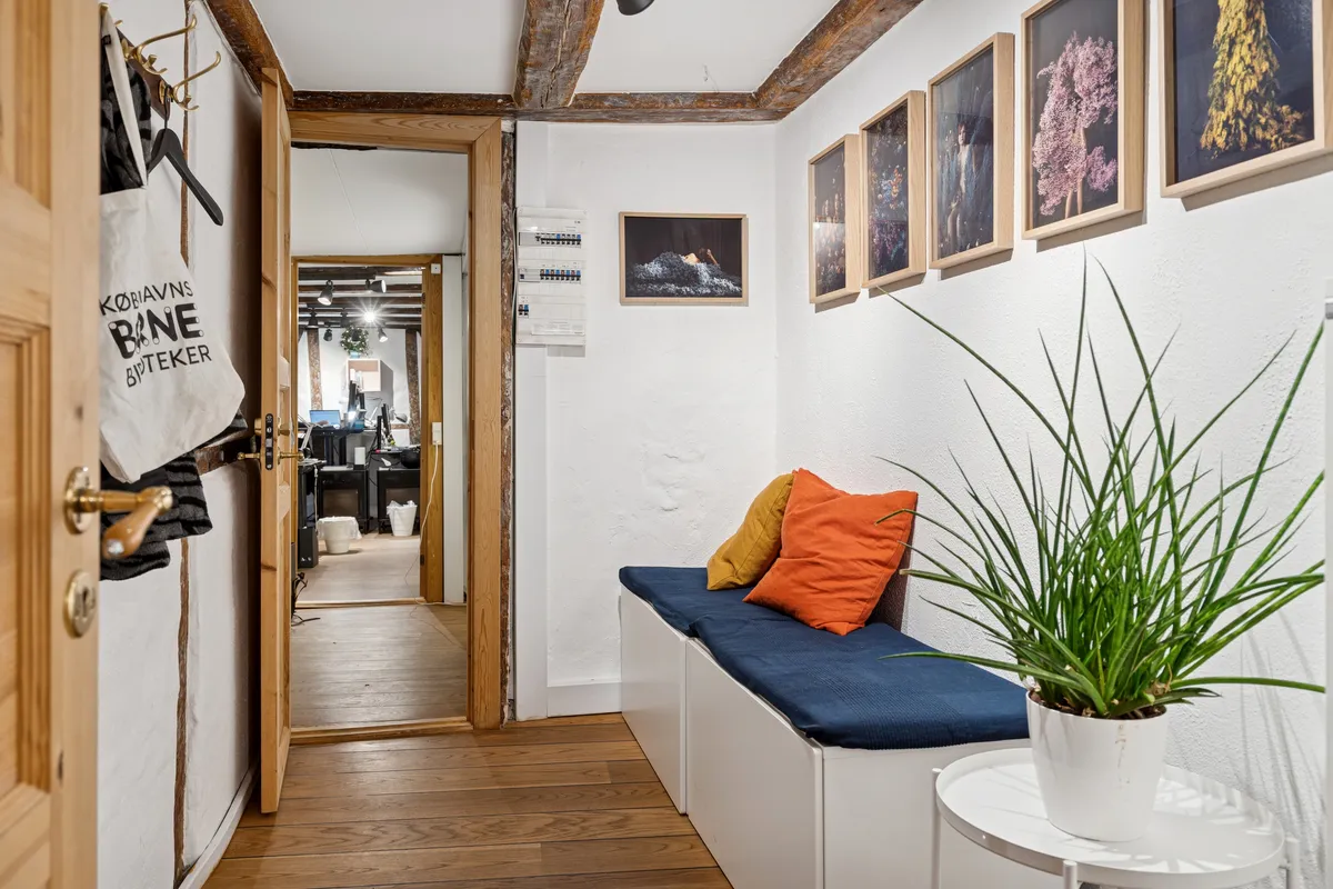 Trægulvet og hvide vægge i kontorrummet med plante og puder. Kontoret har et åbent område med lysekroner og spejl. Der er et bagkuffert og en københavnsbagetaske hængende på væggen.