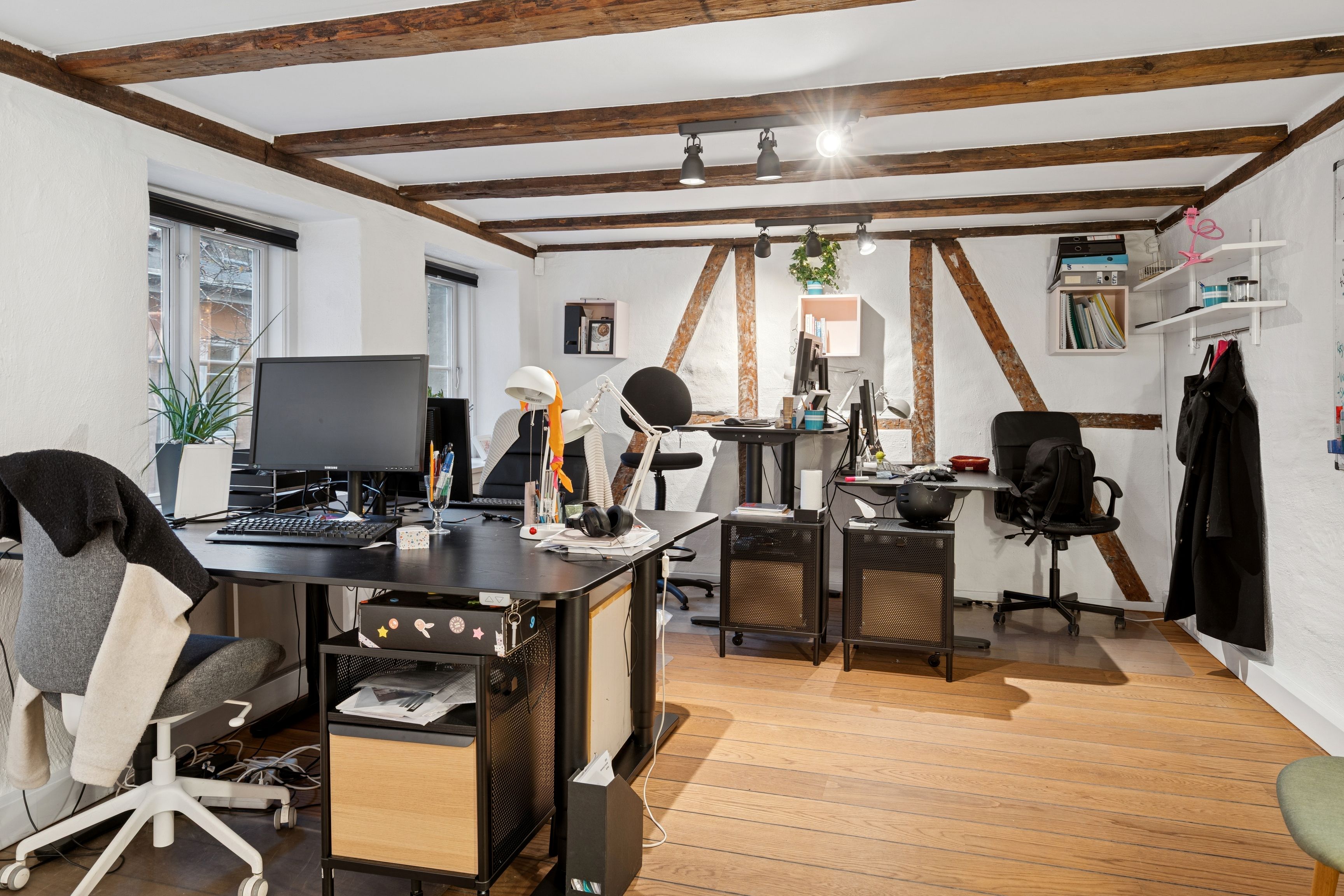 Kontor med computere, skriveborde og lysekroner på loftet, samt hængeklæder og stole i hjørner