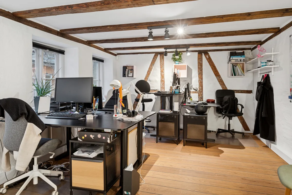 Kontor med computere, skriveborde og lysekroner på loftet, samt hængeklæder og stole i hjørner