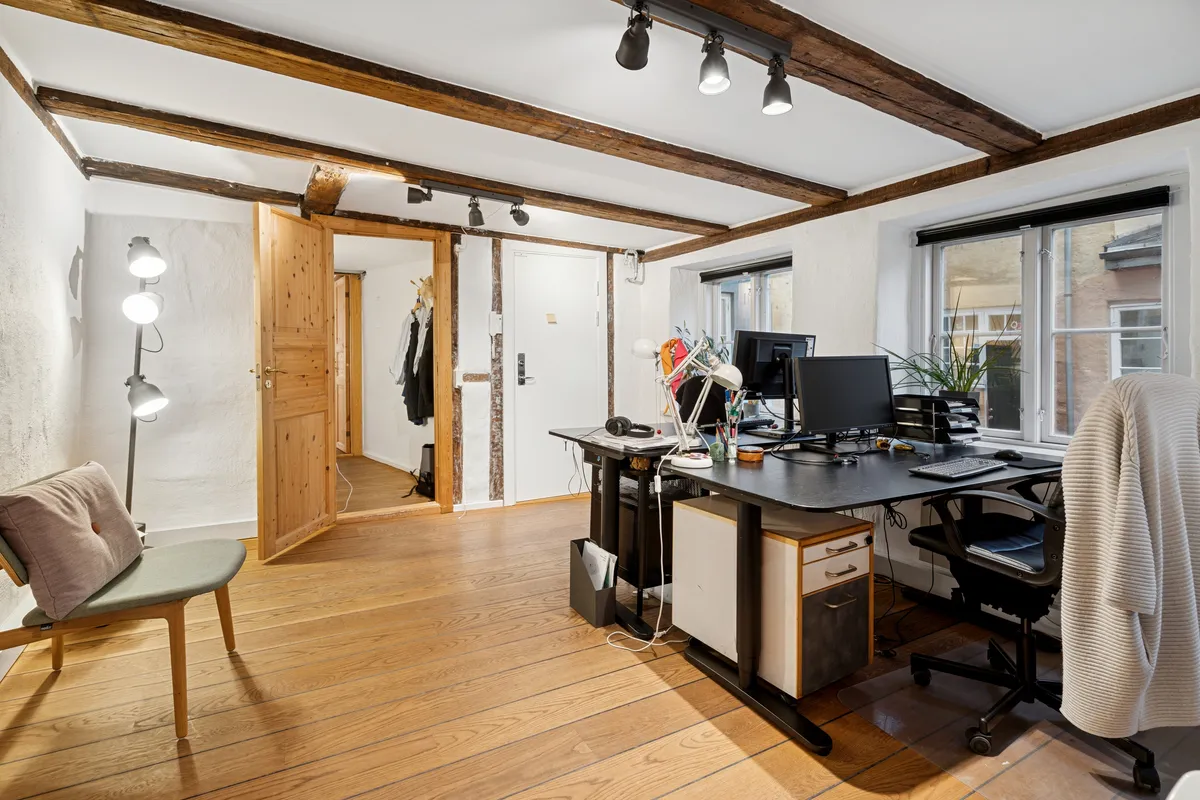 Kontorrum med højt til loftet og lyse træbjælker, skrivebord, to skærme, stol og lampe samt et vinduesvisk.
