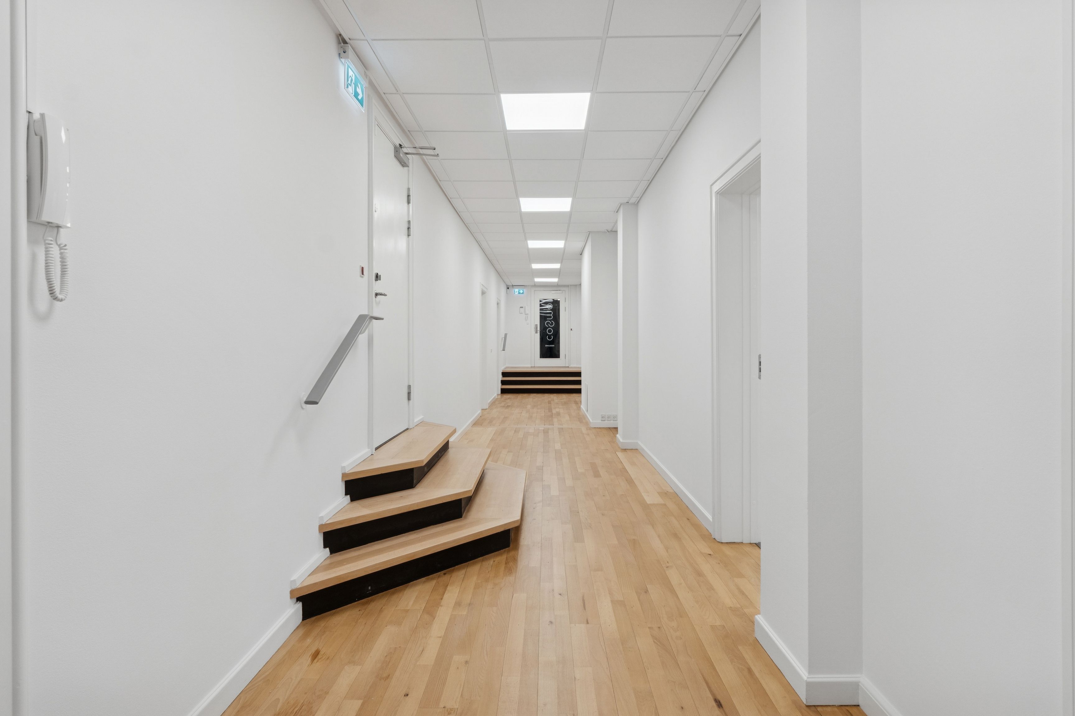 En lys og indbydende kontor med tre trins trætrapper, hvide vægge, moderne loftsbelysning og et hvidt telefonapparat ved siden af en hvid dør på en trægulv