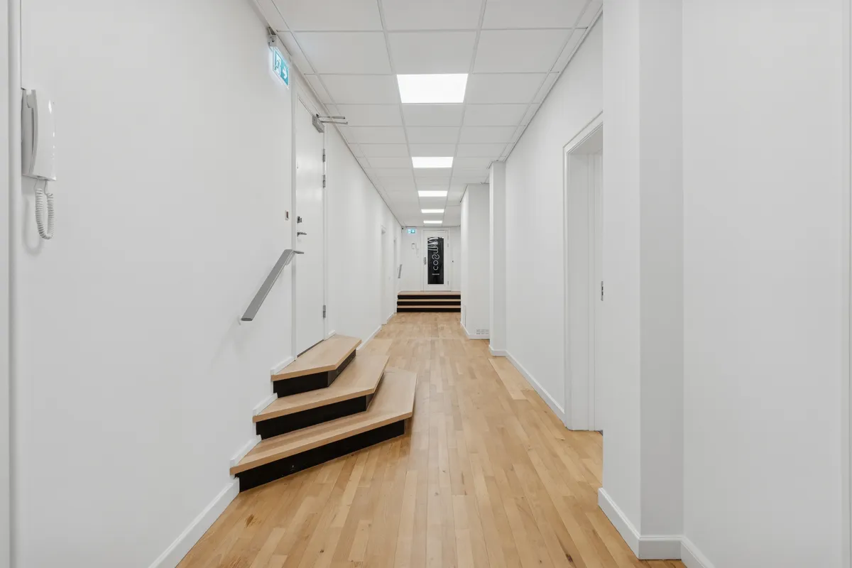 En lys og indbydende kontor med tre trins trætrapper, hvide vægge, moderne loftsbelysning og et hvidt telefonapparat ved siden af en hvid dør på en trægulv