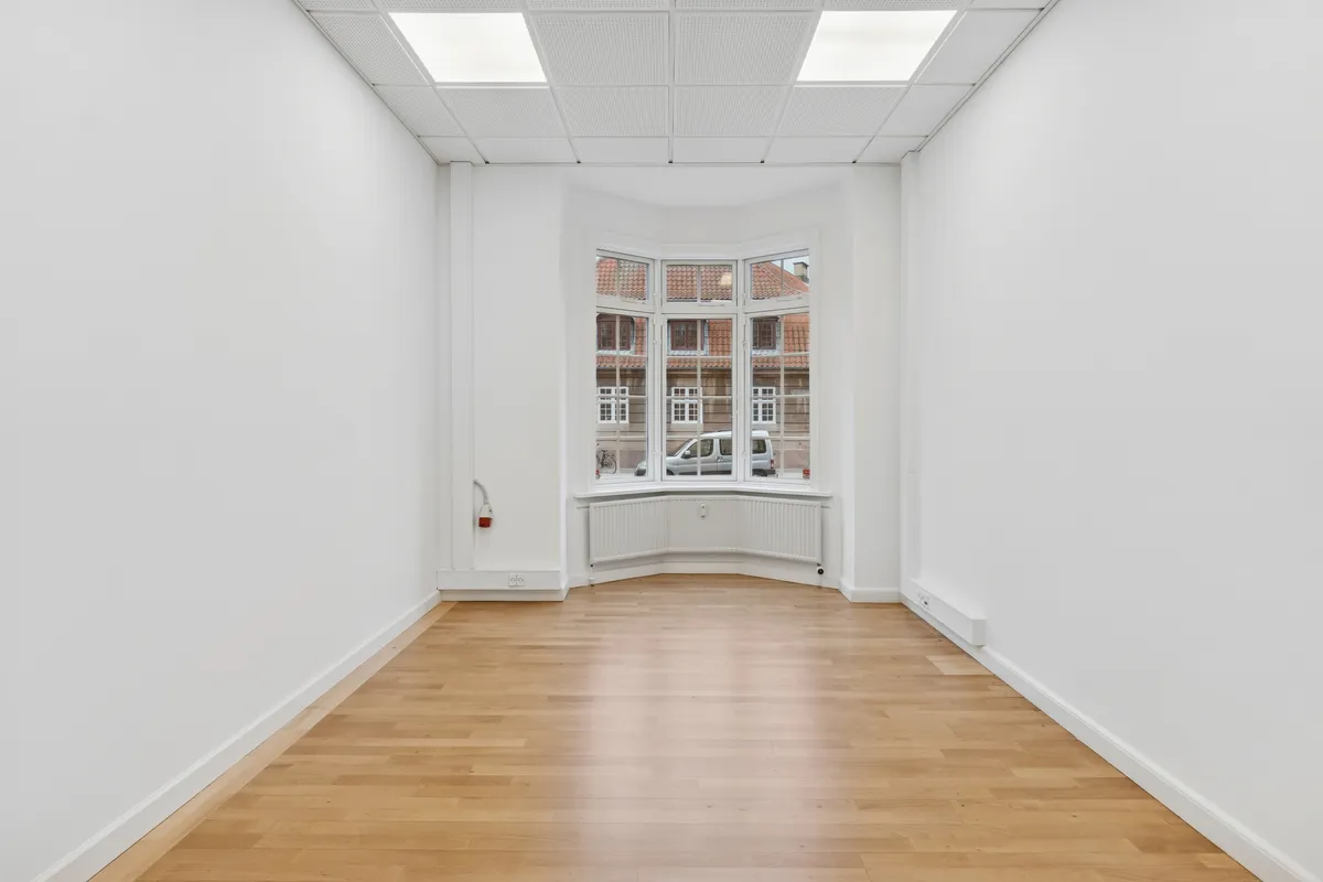 Lyst og indbydende kontor med lyse trægulve og store vinduer til meget naturligt lys. Hjørner med radiatorer og moderne lyskabler for komfortabel arbejdstemperatur.