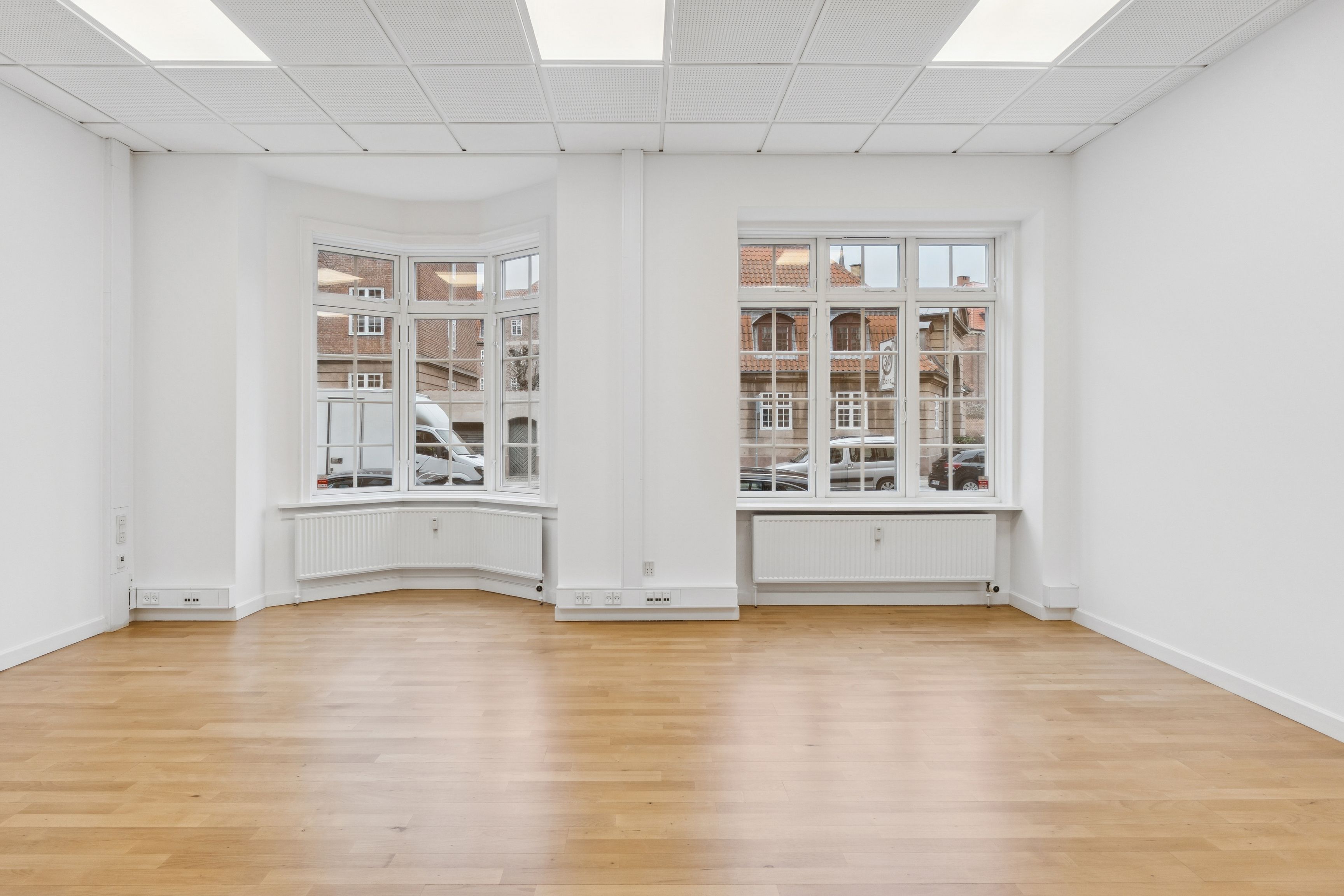 En lys og ren kontor med lysegule gulve og hvide vægge samt store ruder og tavler. To hvide radiatorer er installeret under hver af de to ruder