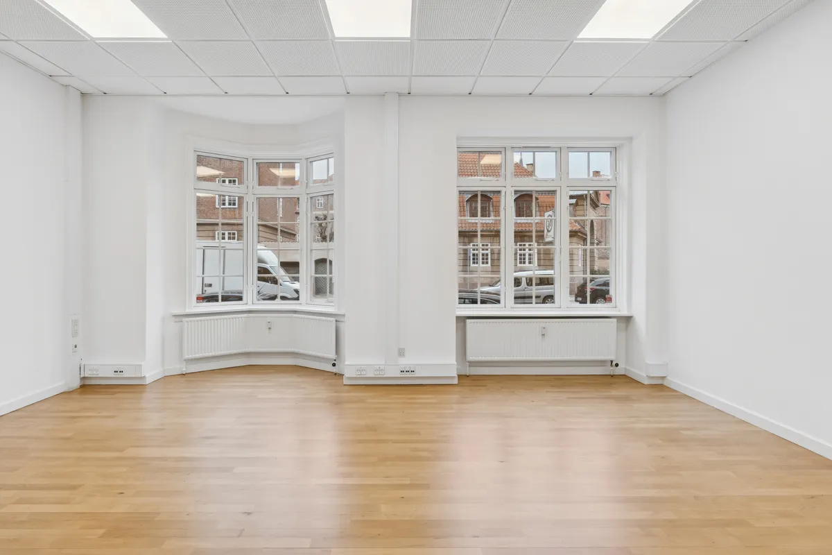 En lys og ren kontor med lysegule gulve og hvide vægge samt store ruder og tavler. To hvide radiatorer er installeret under hver af de to ruder