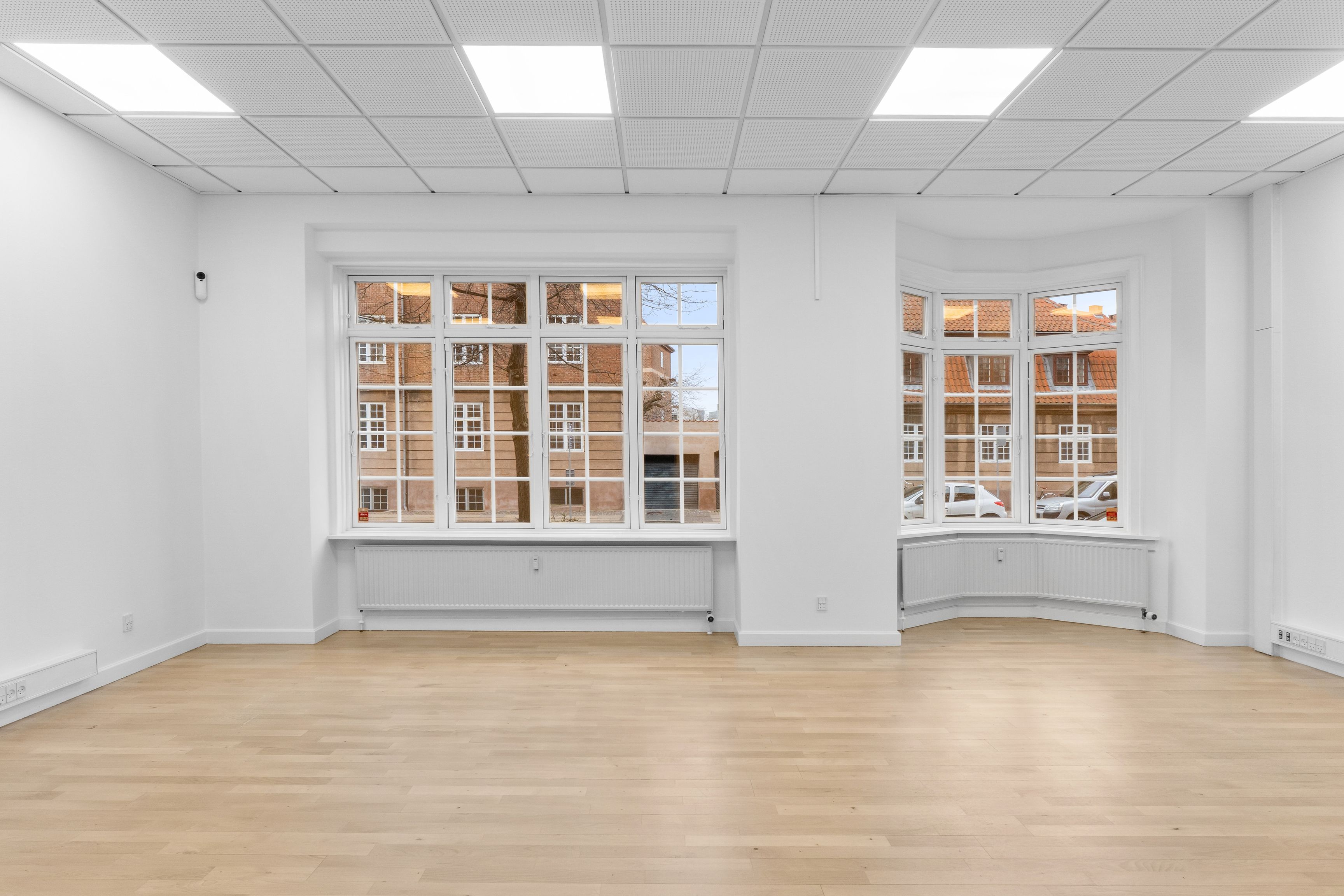 Et renslet kontor med to store vinduer, hvide vægge, moderne gulvbelægning, radiatorer og lyse lofter med lyspaneler. Billedet viser tættere på metrostationen på Fensmarkgade 3 i København N