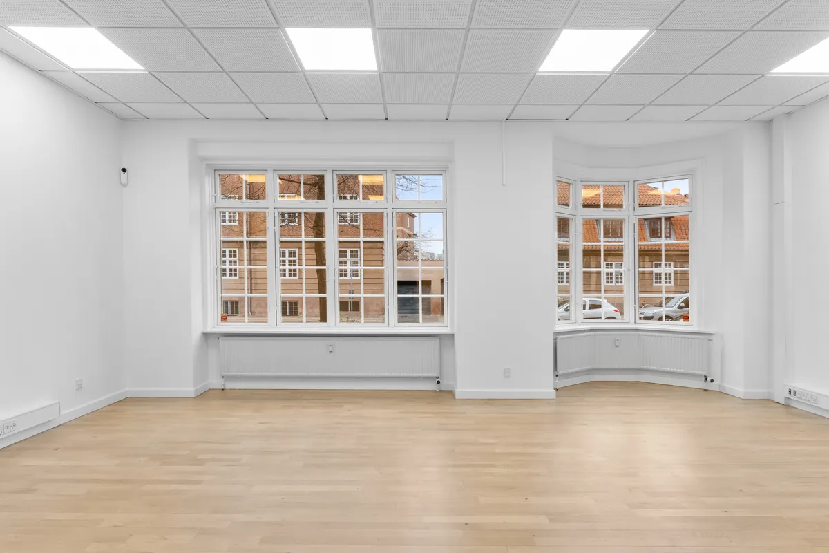 Et renslet kontor med to store vinduer, hvide vægge, moderne gulvbelægning, radiatorer og lyse lofter med lyspaneler. Billedet viser tættere på metrostationen på Fensmarkgade 3 i København N