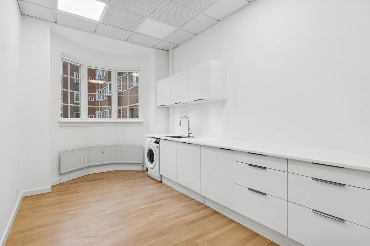 Ren og lys vaskerum med hvide vægge, trægulv, ren og hvidt køkken med underskabe, hvidt loft, en hvid tøjmaskine og et stort bueformet vindue med udsigt til bygninger og gader