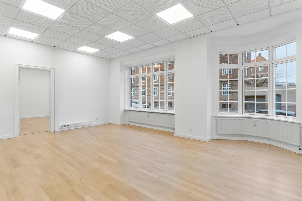 Et lyst kontor med lyse, trægulve og hvide vægge, store vinduer og flere radiatorer. Dørene er hvide og rummet er rent og fuldstændig tomt