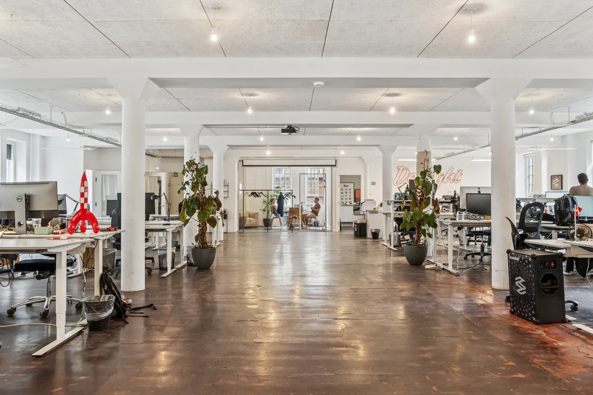 Kontor med moderne møbler og højt loft, store vinduer, lyse loftslamper, en mand i baggrunden og en rødmalet raket på skrivebordet