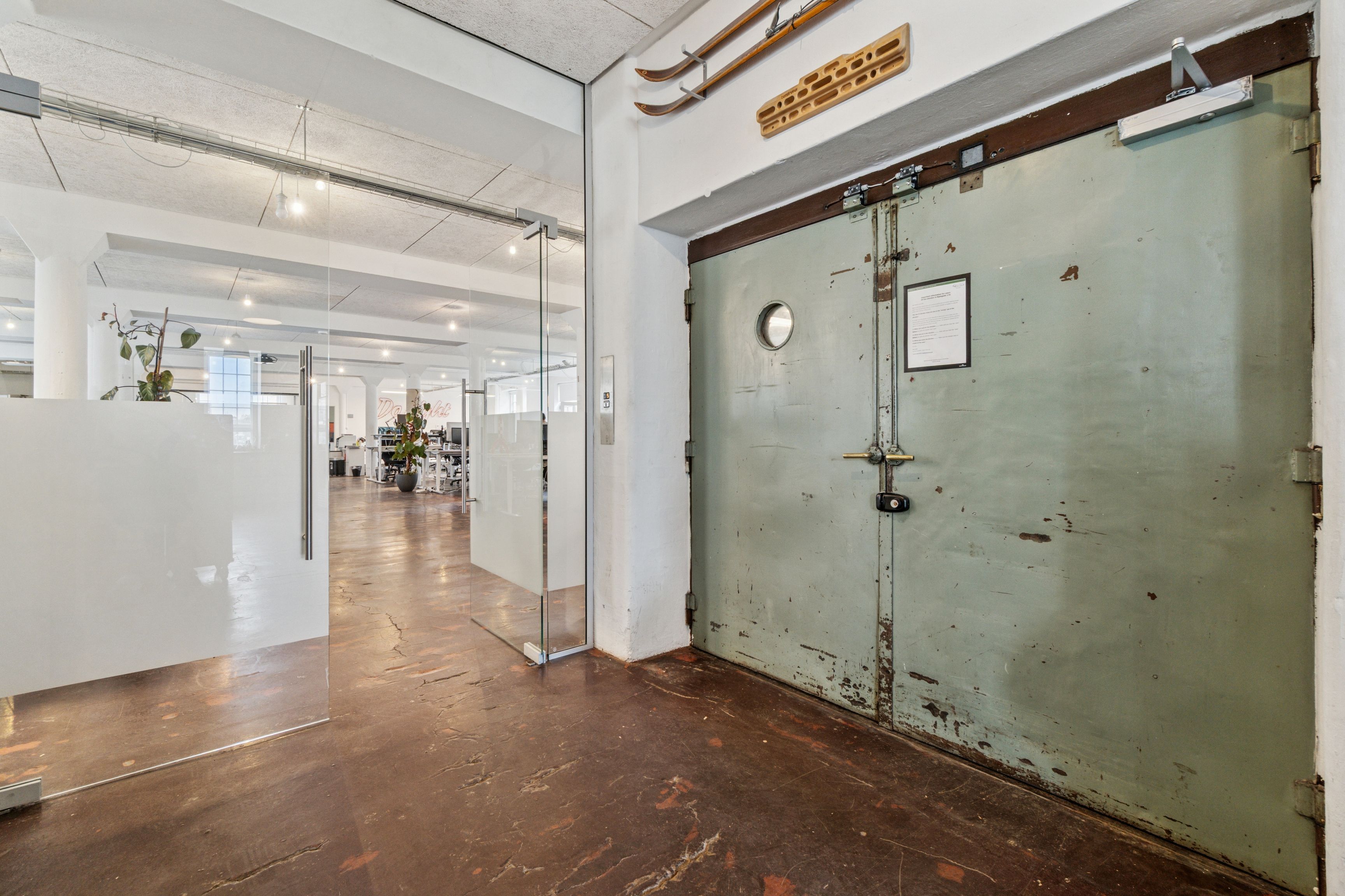 Kontorrum med lyse glasdøre og gammel hoveddør, indvendige høje lofter og lysepunkter, toppe med ski, foran et skrivebord med plante og metrostation i baggrunden