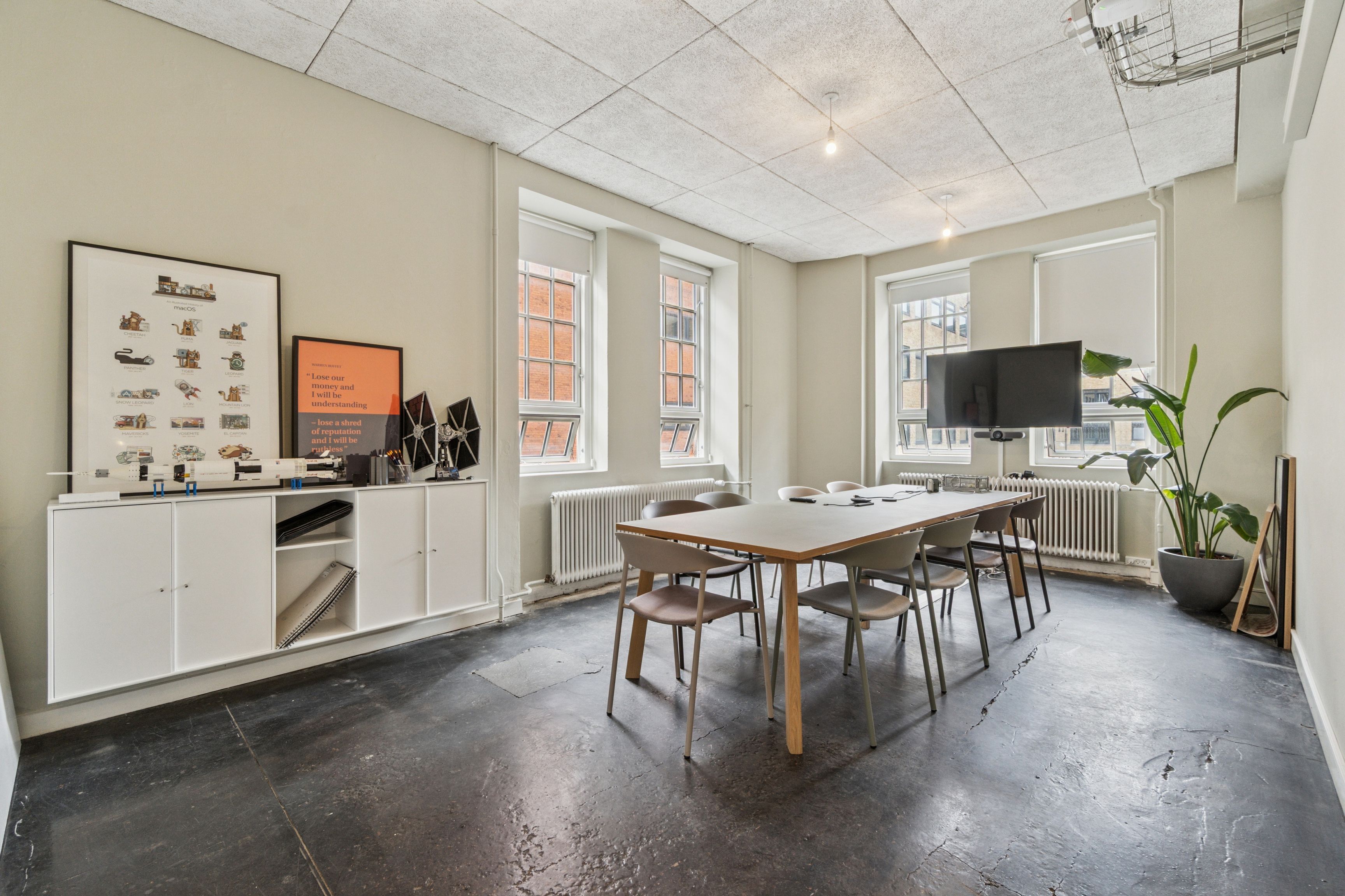 Kontorrum med højt til loftet, moderne møbler, en stor tv-skærm og vinduer med udsigt til byen, samt en stor grøn plante i en grå potte