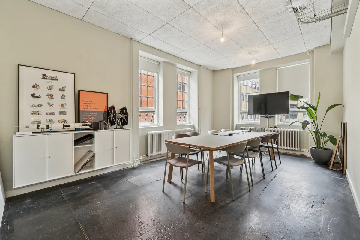 Kontorrum med højt til loftet, moderne møbler, en stor tv-skærm og vinduer med udsigt til byen, samt en stor grøn plante i en grå potte