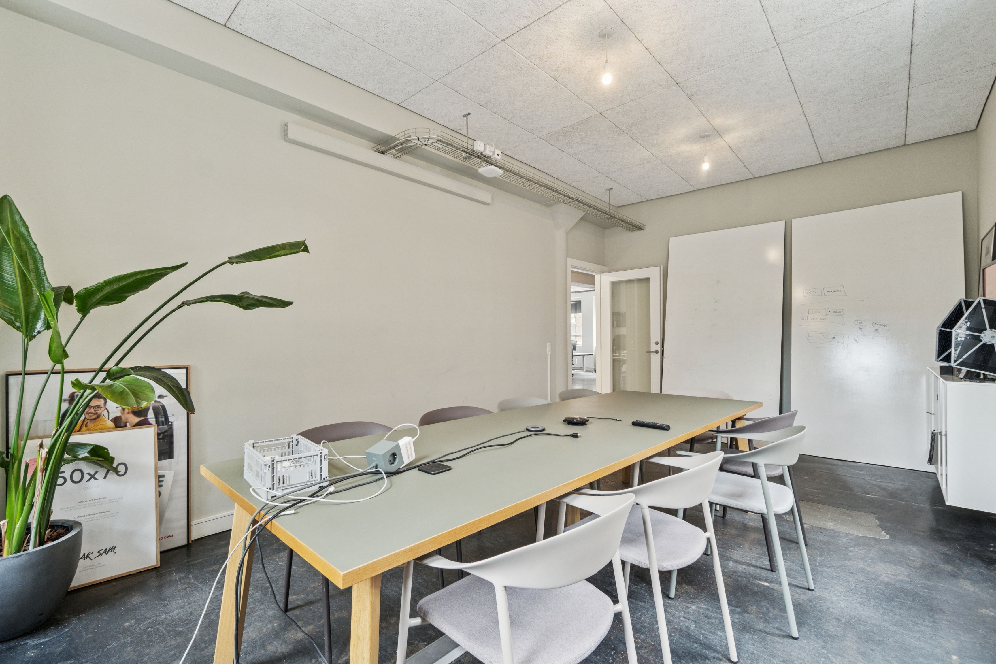 Kontorrum med hvide vægge, lyseloft, høj gulvdekning og moderne møbler. Værelset er delt op med en lang, gul tabel med tilsluttede kabler. Højre side har to hvide vægmonterede tavler.