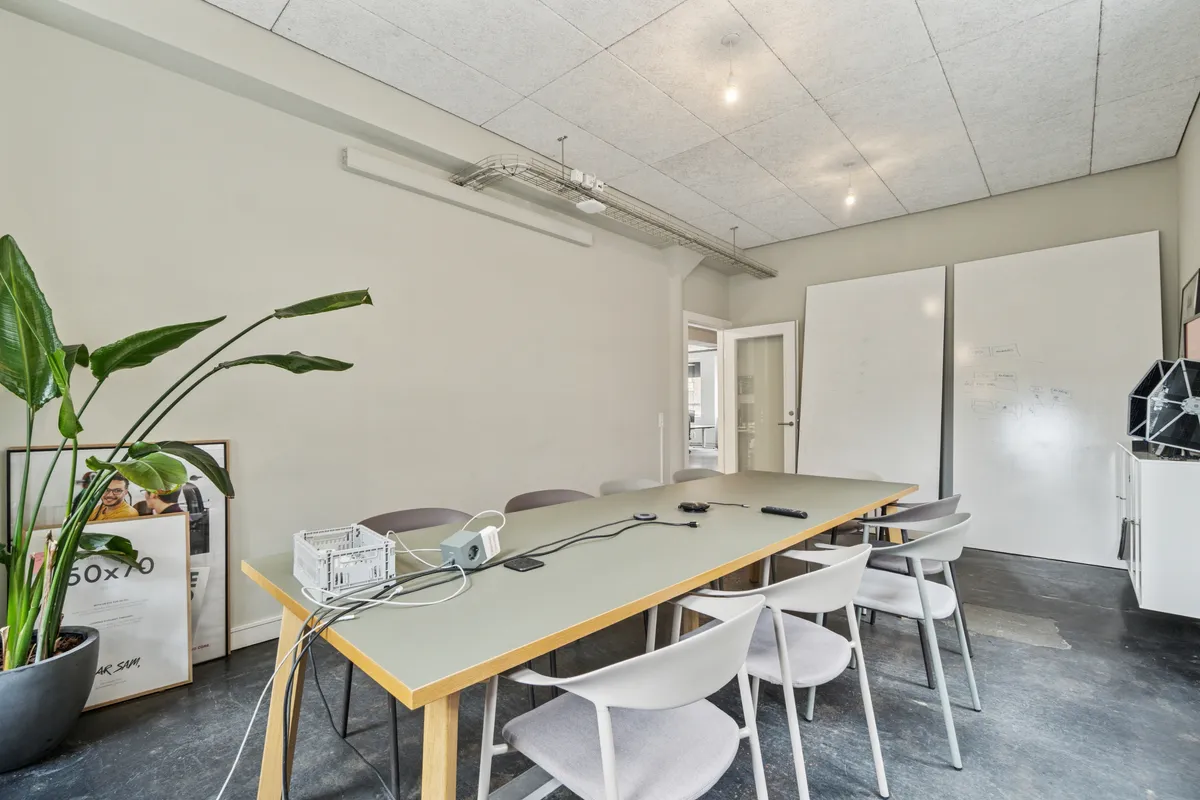 Kontorrum med hvide vægge, lyseloft, høj gulvdekning og moderne møbler. Værelset er delt op med en lang, gul tabel med tilsluttede kabler. Højre side har to hvide vægmonterede tavler.