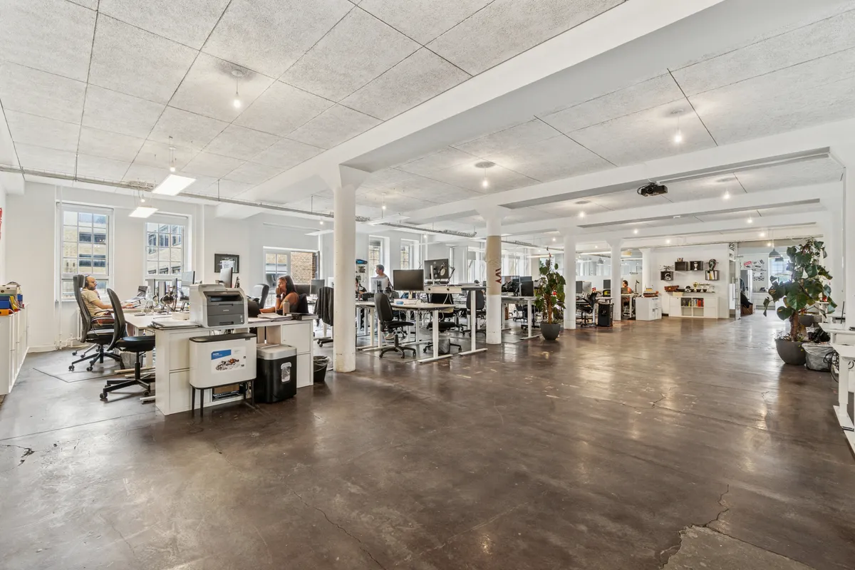 Kontorrum med store vinduer, højt loft og masser af lys. Kontorbord, skrivemaskine, plante og mennesker. Beliggenhed tæt ved metrostation.