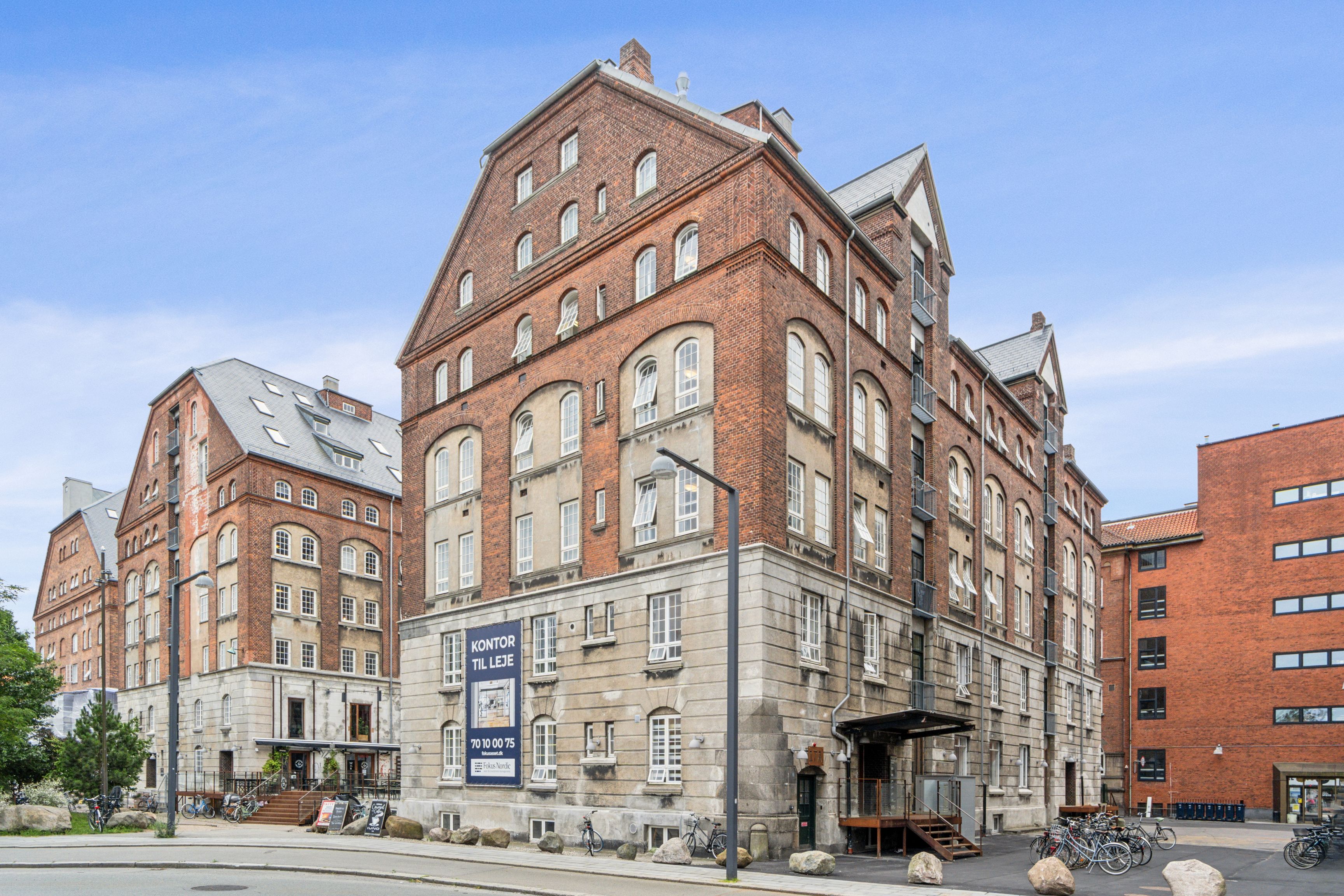 Rå og lyse kontorlejlighed på Islands Brygge med skiltet "Kontor til leje", der vender ud mod gågaden med mange cykler og grønne planter