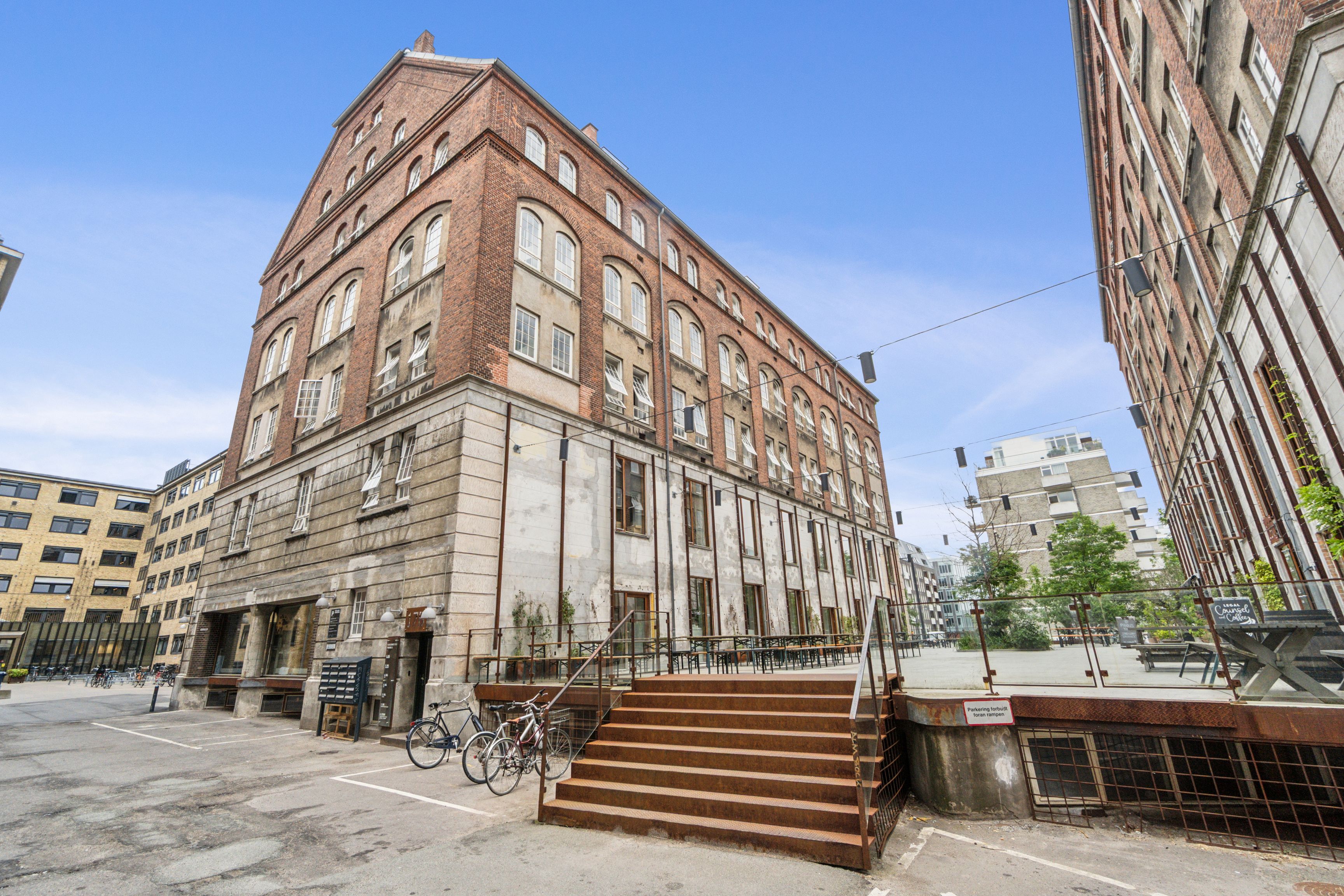 To cykler er parkeret uden for kontoret med røde trin, der fører op til døren og terrassen med bænke og bord. Bygningen har røde mursten og lyseruder, og det er en solrig dag.