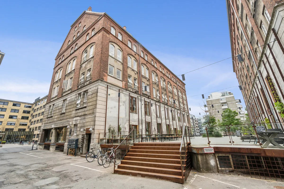 To cykler er parkeret uden for kontoret med røde trin, der fører op til døren og terrassen med bænke og bord. Bygningen har røde mursten og lyseruder, og det er en solrig dag.