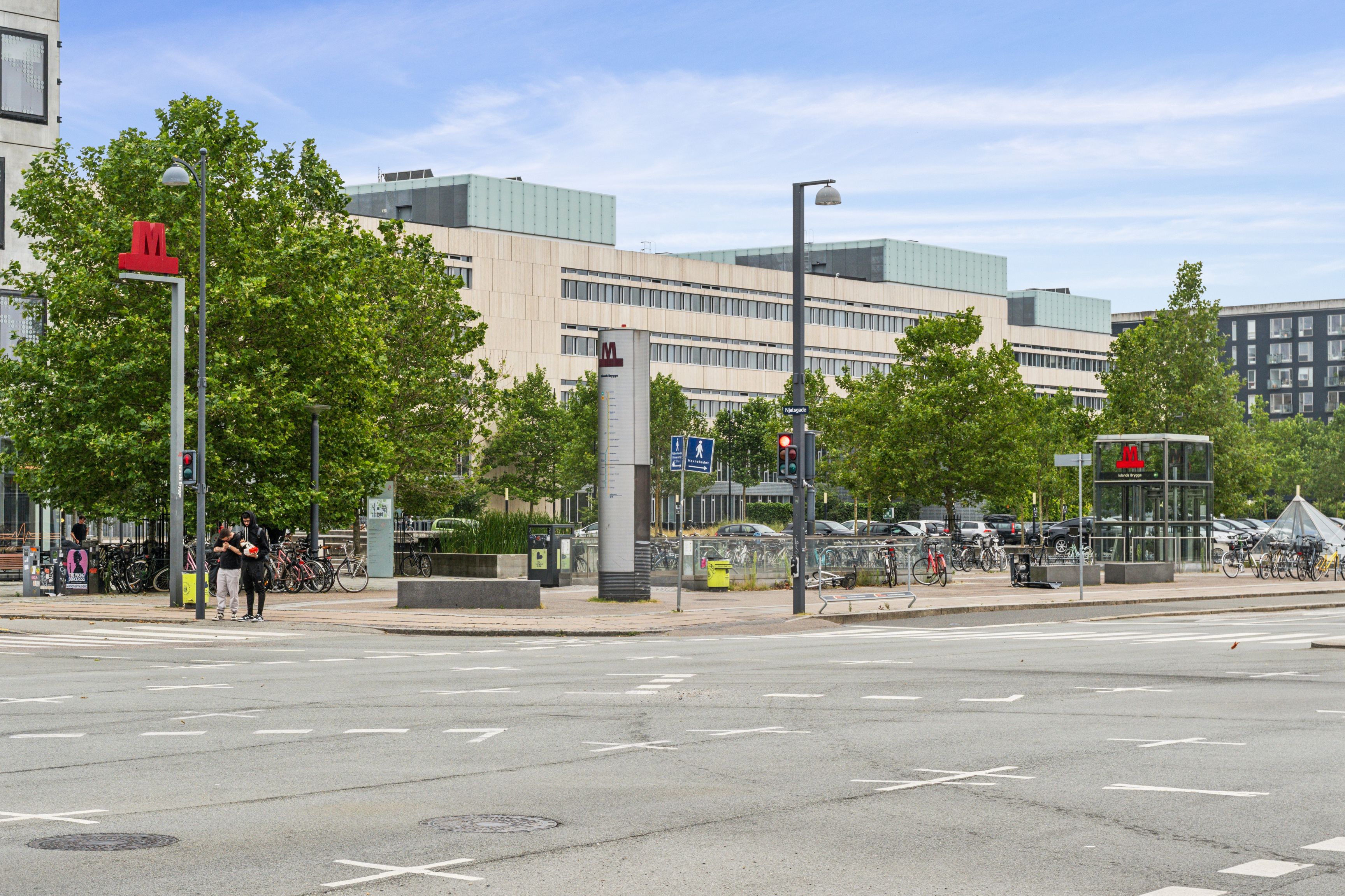 To mennesker står ved en stor bygning og snakker, mens der er flere parkerede biler og cykler i baggrunden