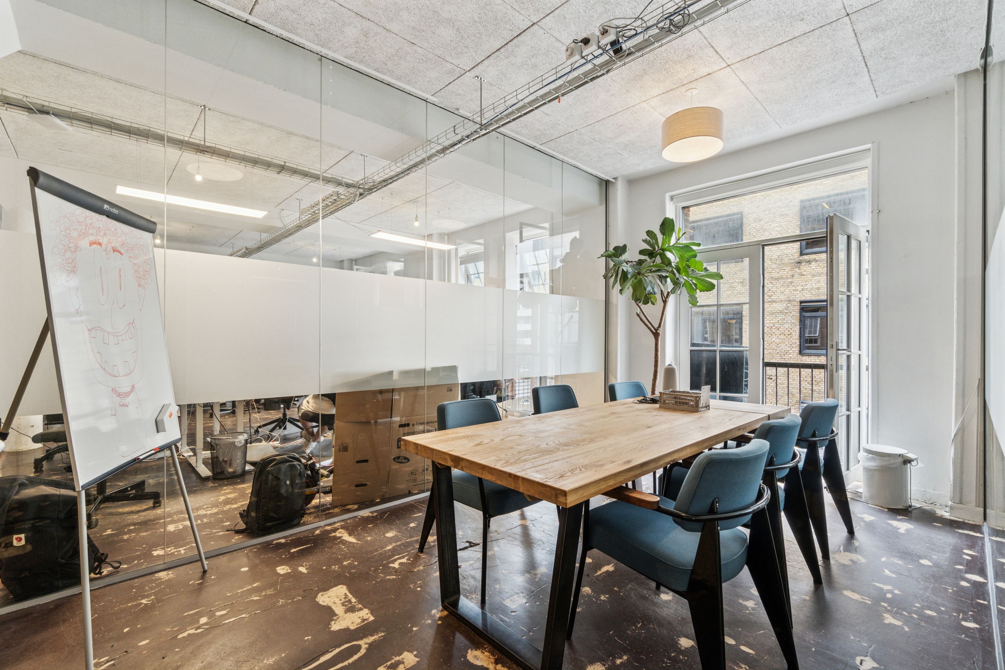 Kontor med mødebord og lyse buer, lyse hvide vægge og glasvinduer, hvor et whiteboard med tegning står. Plante, lysekrone og åbne vinduer med udsigt til byggede bygninger