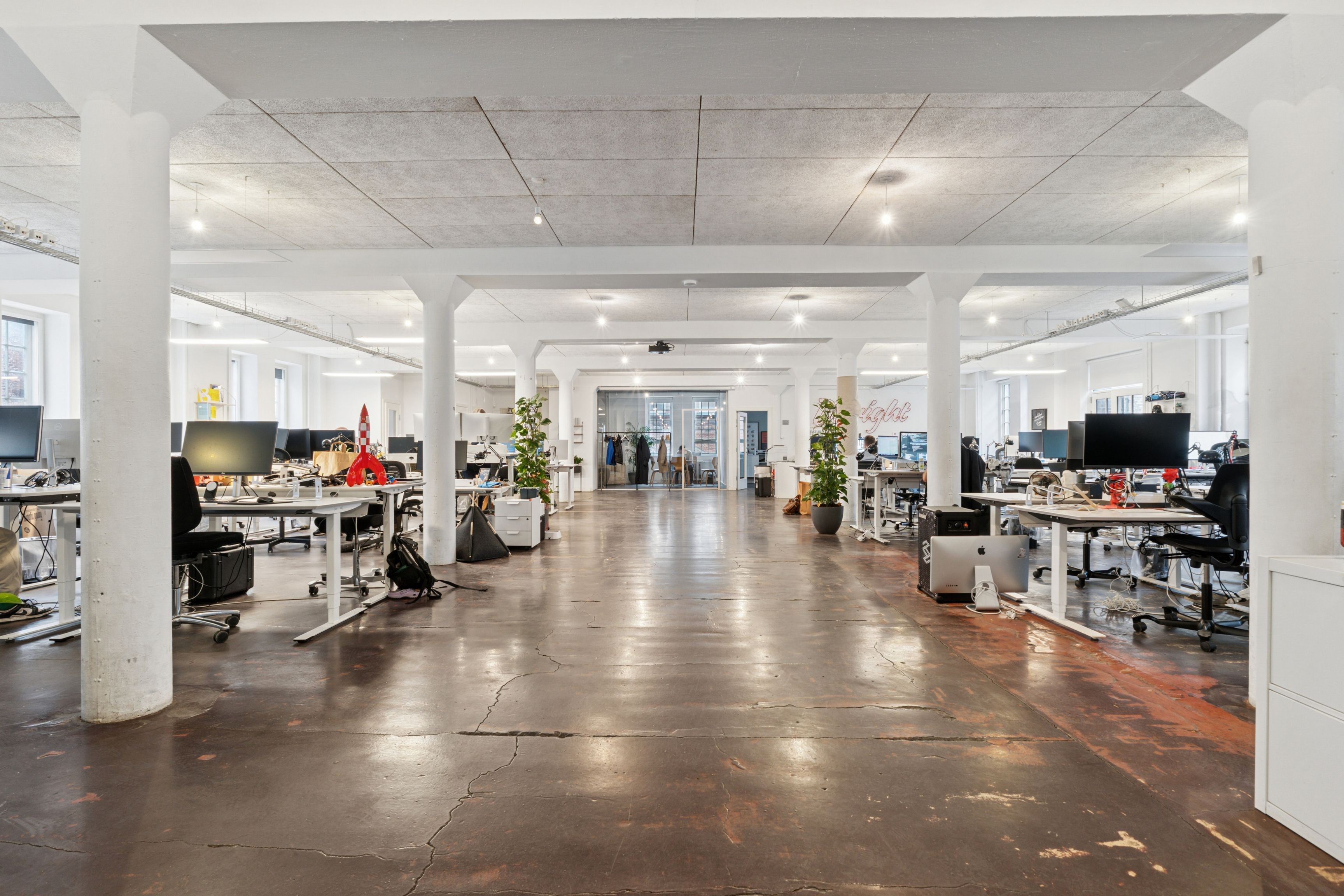 Kontorrum med lyse lofter, hvide søjler, moderne møbler, computere og plantepoter placeret i tæt bebygget område med udgang til gadebaner