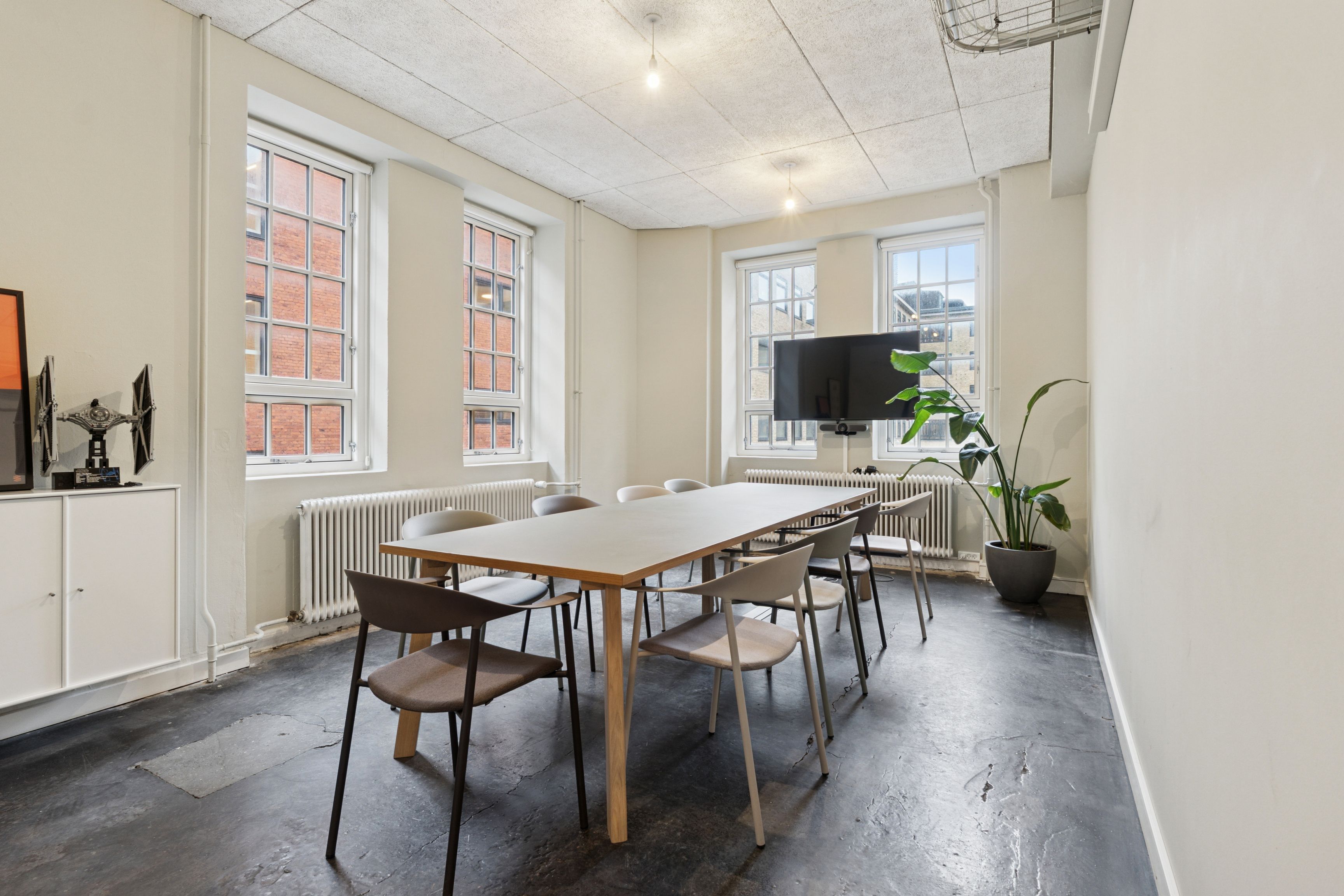 Minimalistisk mødelokale med lyse ruder, hvidt interiør og moderne møbler, højt til loftet, et træbord, tv og grønne planter samt lysekroner i loftet