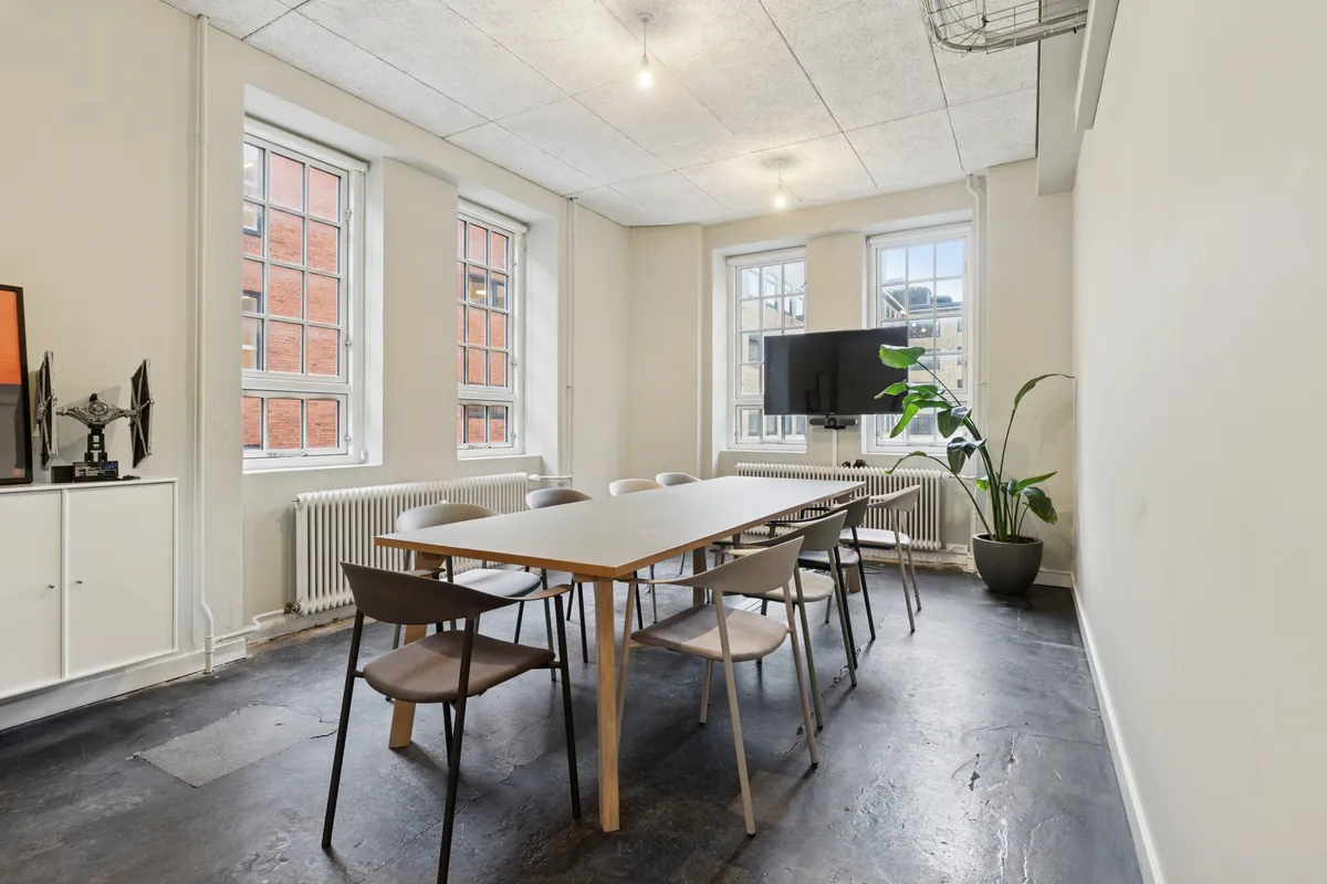 Minimalistisk mødelokale med lyse ruder, hvidt interiør og moderne møbler, højt til loftet, et træbord, tv og grønne planter samt lysekroner i loftet