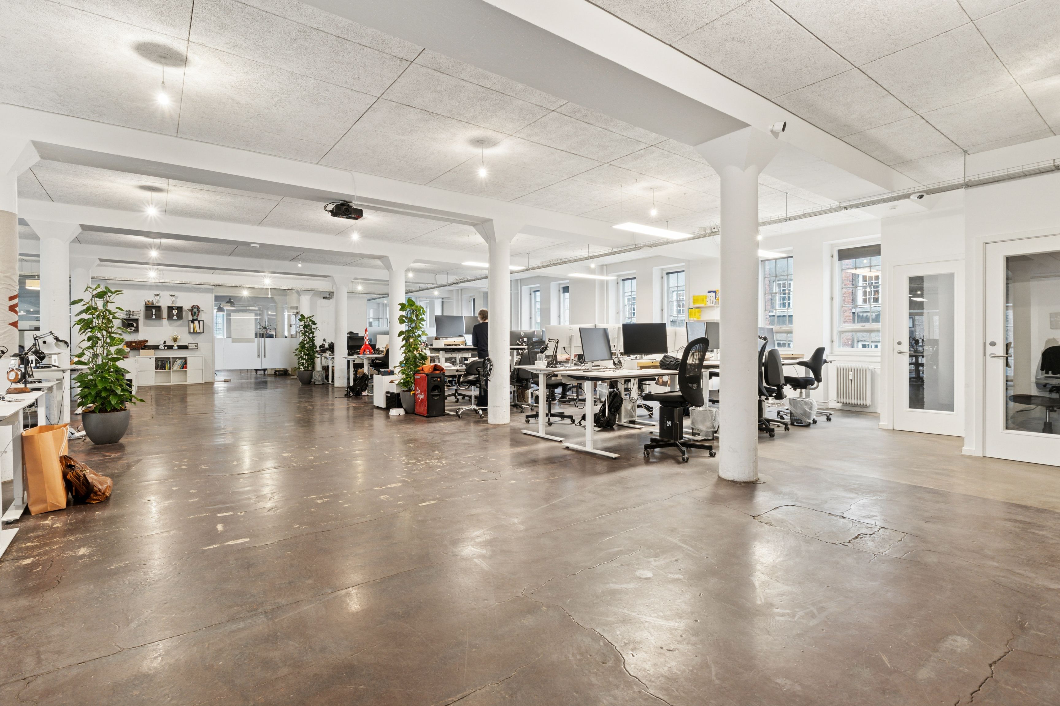 Kontorlokaler med store vinduer, hvide vægge og lyse lofter, skriveborde og stole, en plante og en person, som står ved et skrivebord, samt teknologi som computere og projektorer