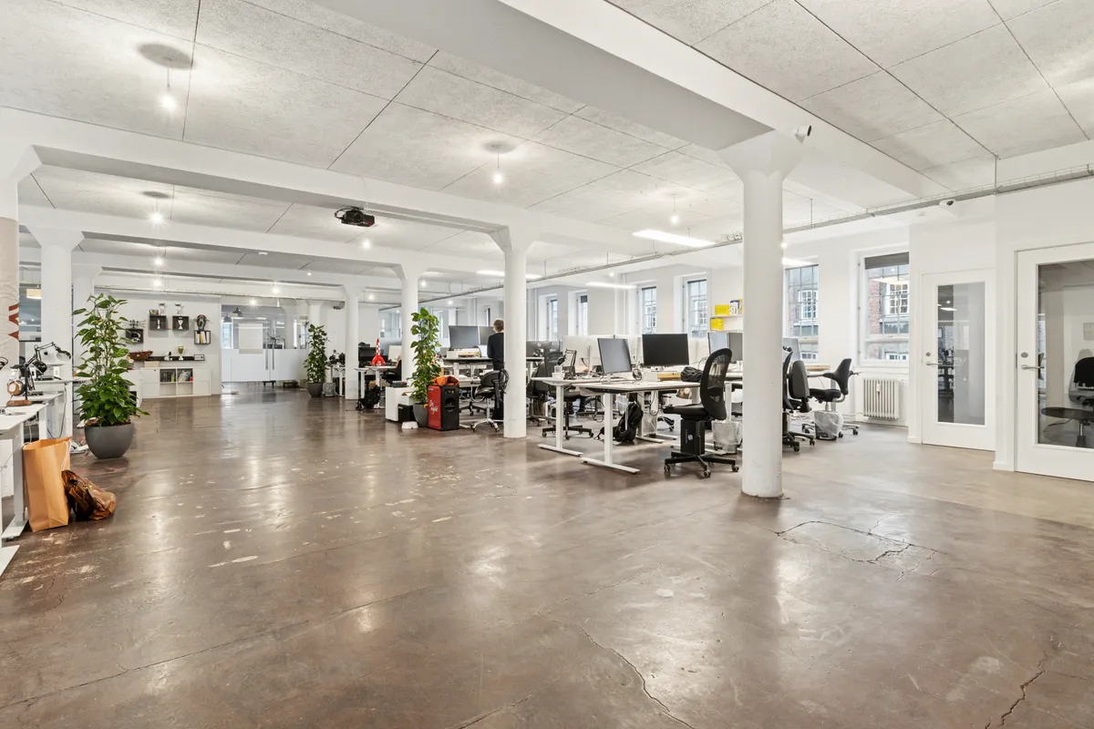 Kontorlokaler med store vinduer, hvide vægge og lyse lofter, skriveborde og stole, en plante og en person, som står ved et skrivebord, samt teknologi som computere og projektorer
