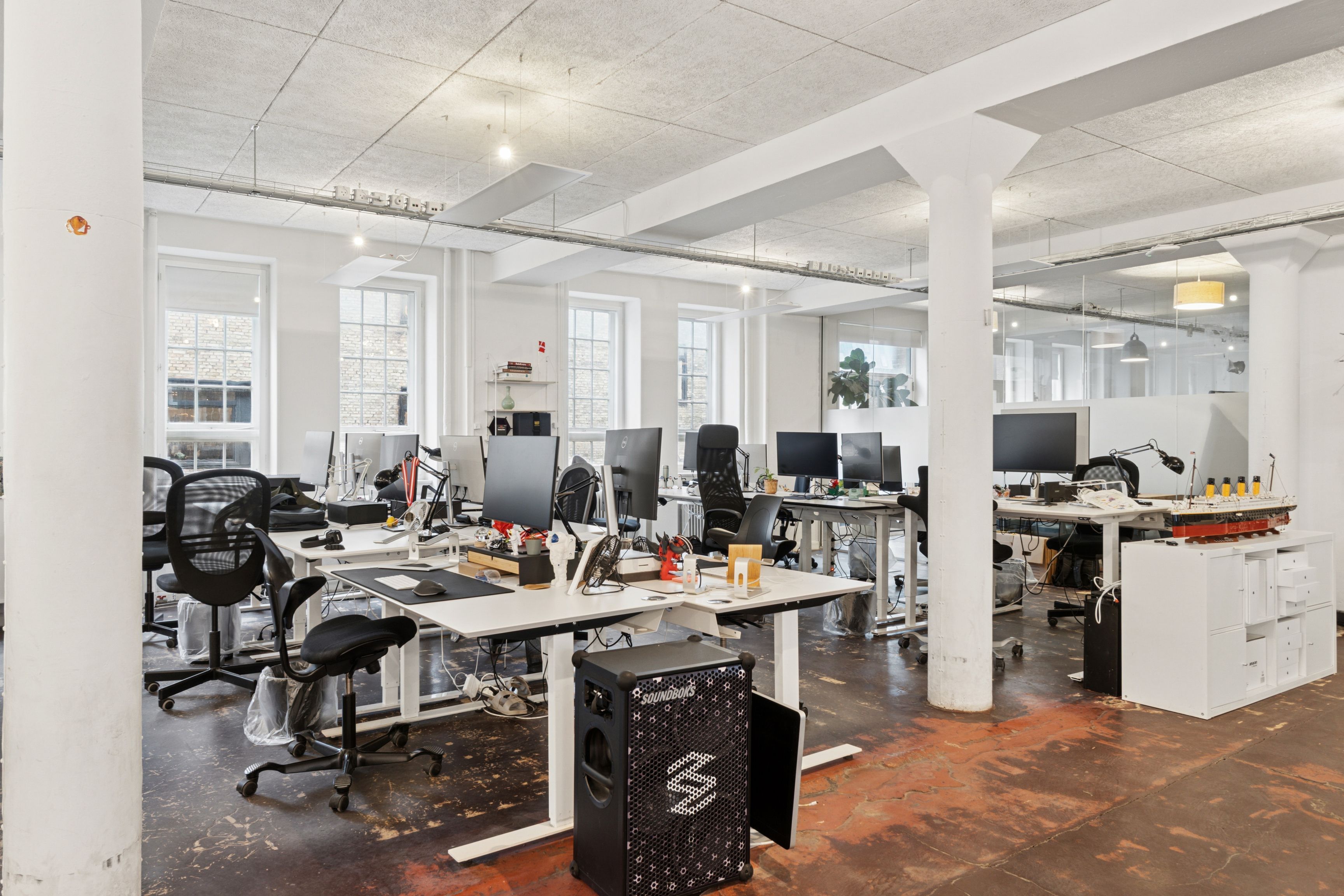 Kontor med højloft, lysevinduer og moderne møbler. Trægulv og lyse loftslampe. Tastaturer, skærme og bærbare computere. Højtalere og modelbåd på skrivebord.