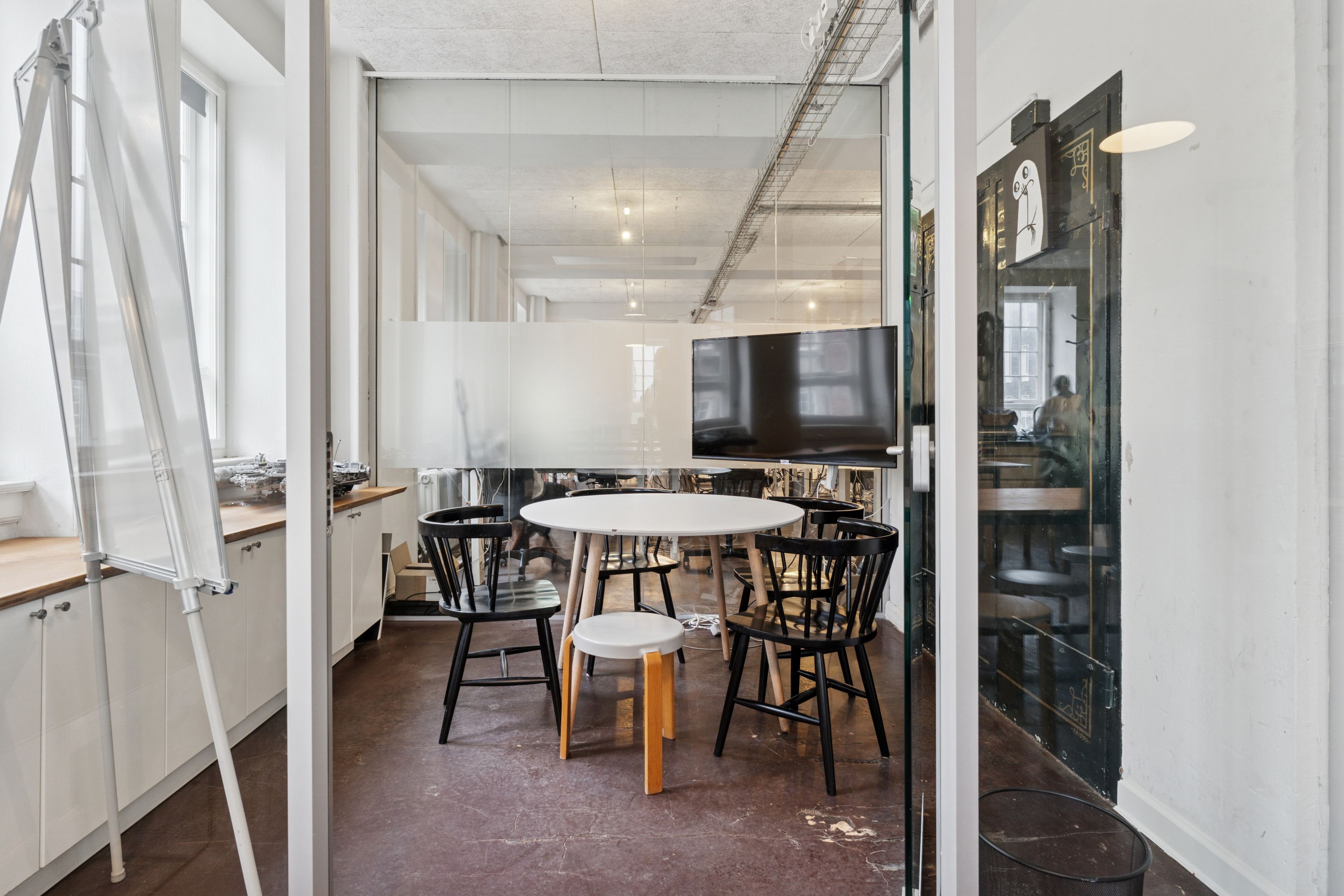 Kontorrum med glasvægge og moderne møbler, hvor der er en hvid rund bord med stol og en stor sort tv-skærm. Der er et skrivebræt med en hvid stol og et skraldspand.