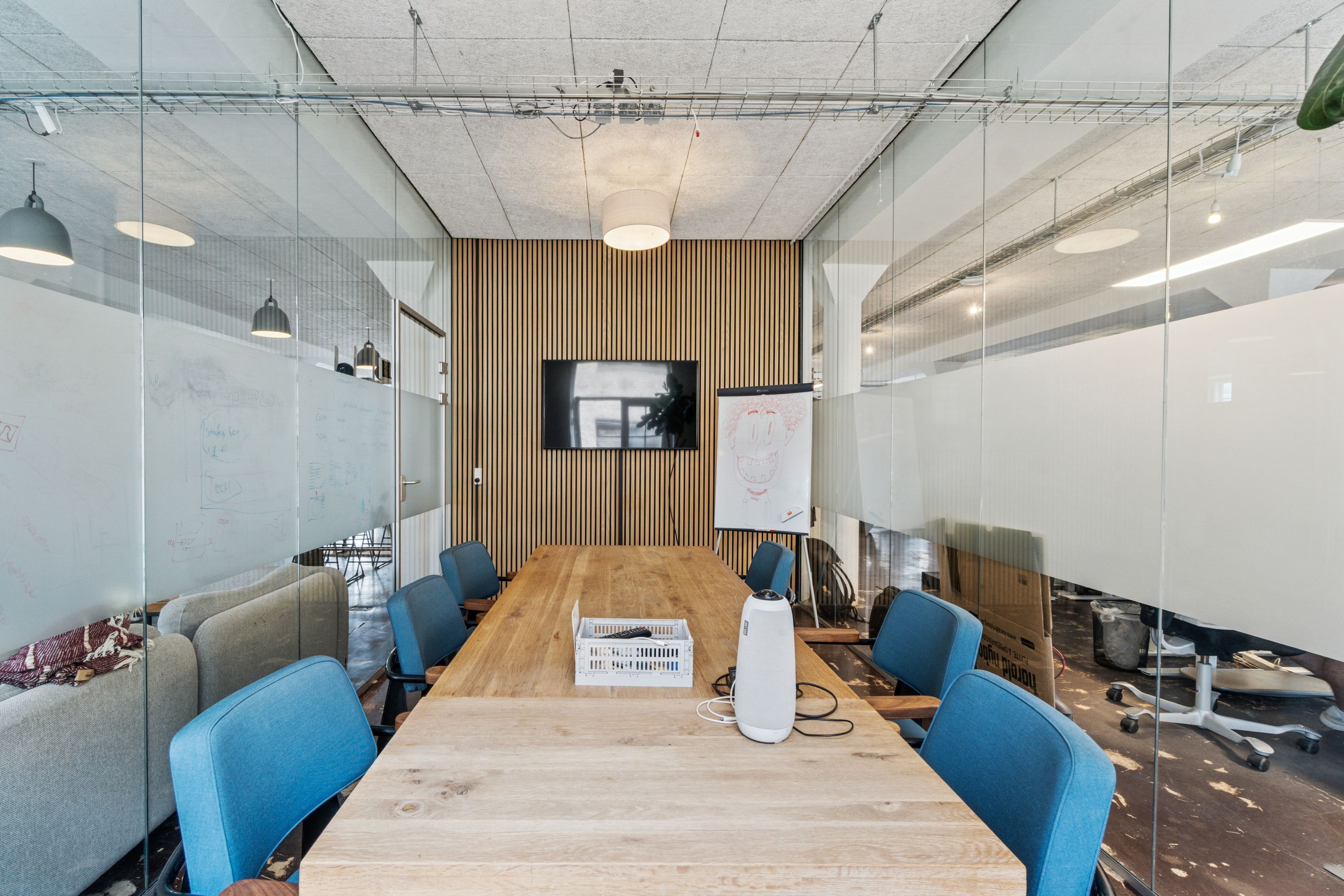Kontorrum med mødebord, hvid tavle og lyse loftslamper, blå stole og sætte trægulve i en stor stue med glasvægge