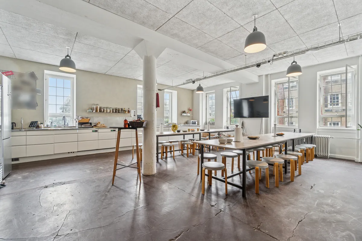 Kontor med langt bord og stole i loftstil. Væg med vinduer, hængelampe, køleskab og en tv-skærm. Hjørne med køkken med håndvask og apparater. Vægge med højde og simpel indretning.