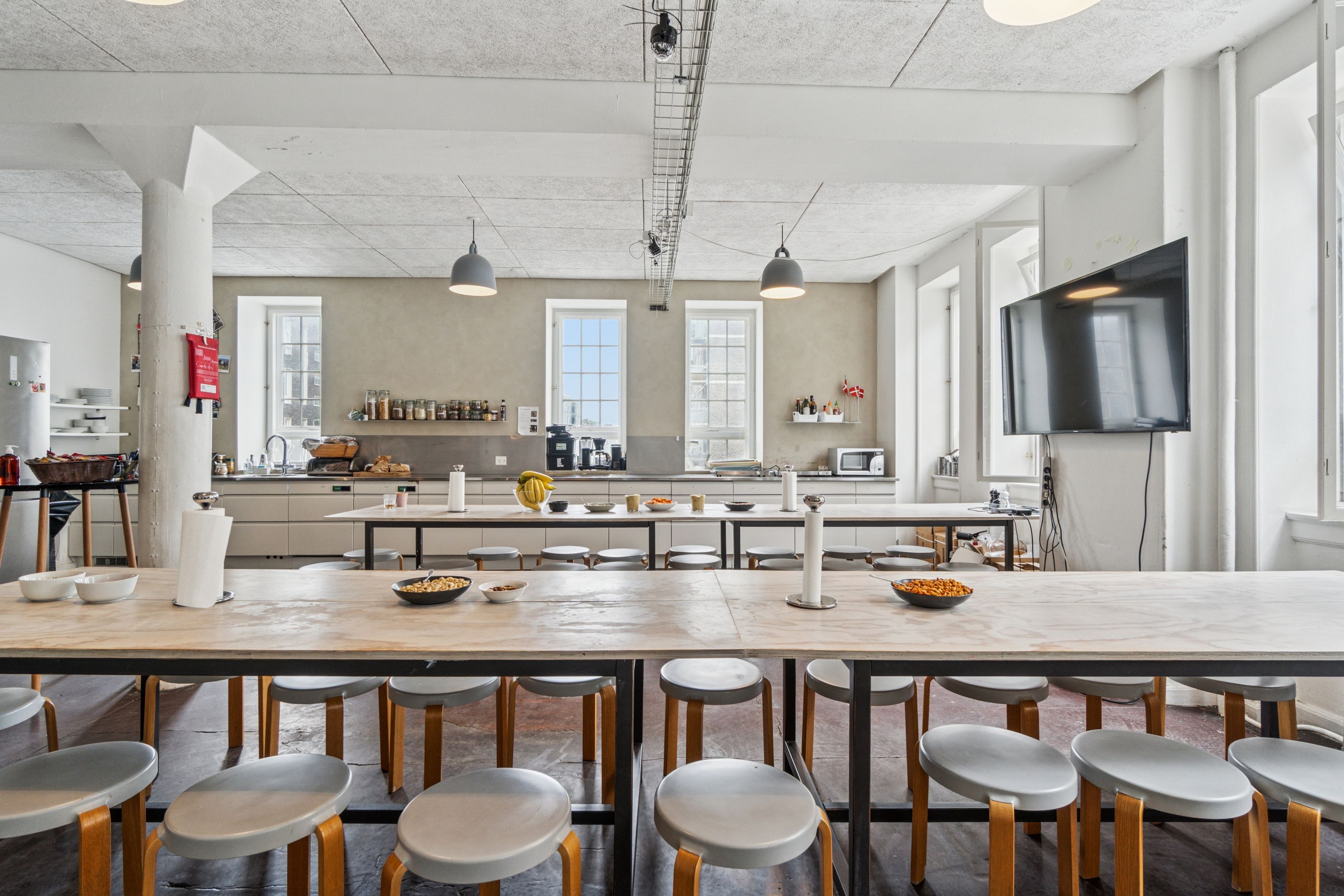 En stor og lys køkkenlokale med langt træbord og høje stole samt flere skuffer og apparater i baggrunden og flere lamper i loftet
