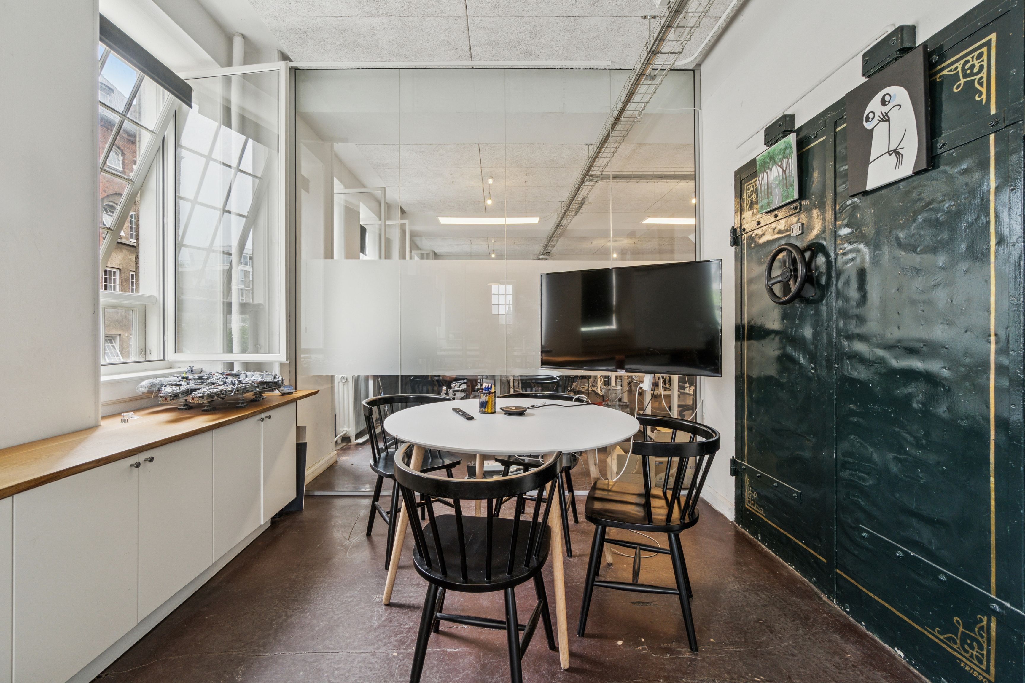 Kontor med masser af lys, hvide vægge og møbler, stort tv, to stolpemonterede vinduer og særligt dørblad på kant.