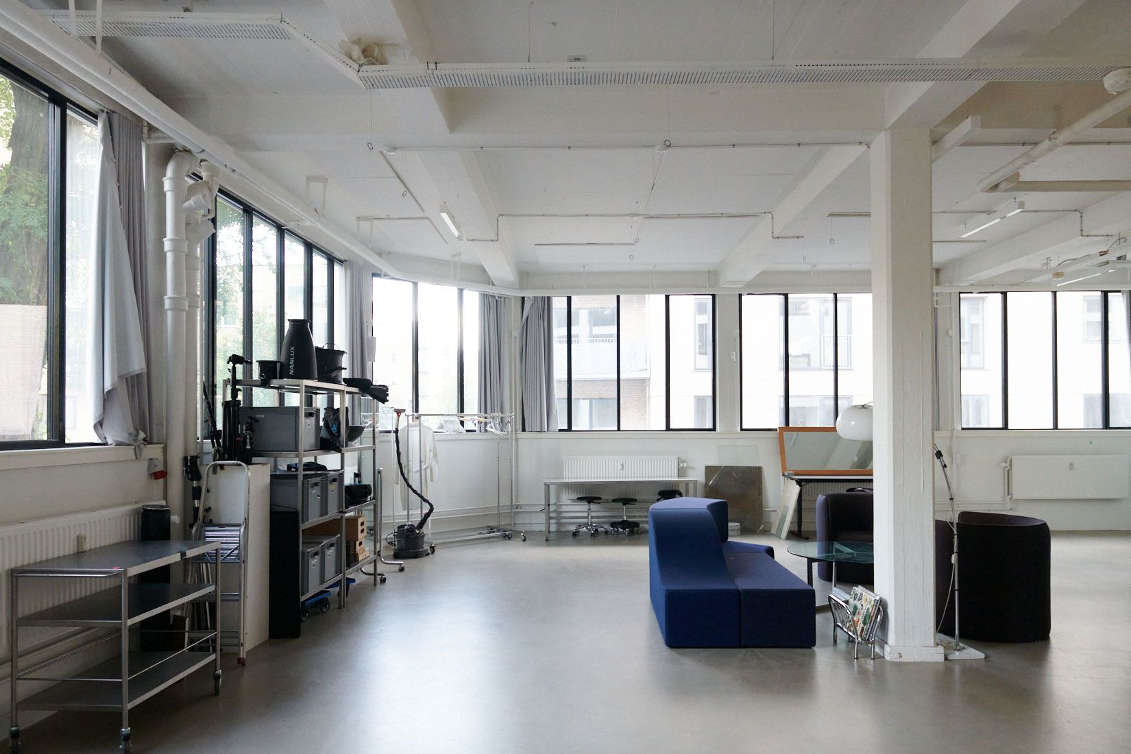 Kontorlejemål med stort vindue, store højlofter, moderne møbler og glasbord, ståbord, sofa og kaffemaskine samt radiatorer og hvide vægge