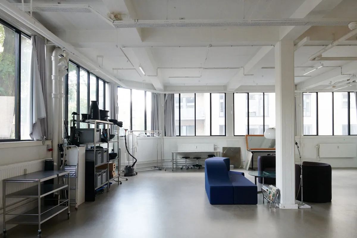 Kontorlejemål med stort vindue, store højlofter, moderne møbler og glasbord, ståbord, sofa og kaffemaskine samt radiatorer og hvide vægge