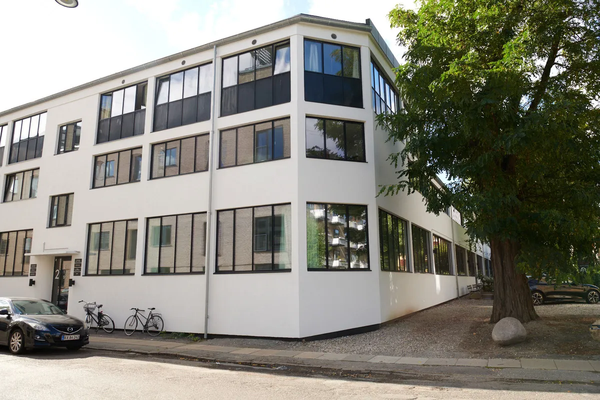 Kontorbyggeri med store ruder og to cykler udenfor, et parkeringsområde med en stor sten og træ, en bil og en lyseblå Mazda bil parkeret på siden