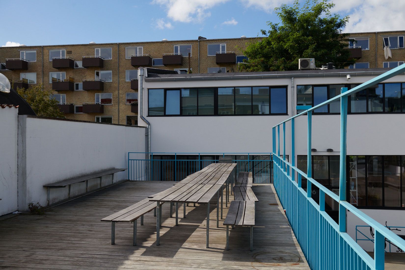 Kontorlejemål med grønne områder og flere transportmuligheder i gåafstand. Forlagshuset med lyse vinduer og terrasseområde med bænke og spisebord.