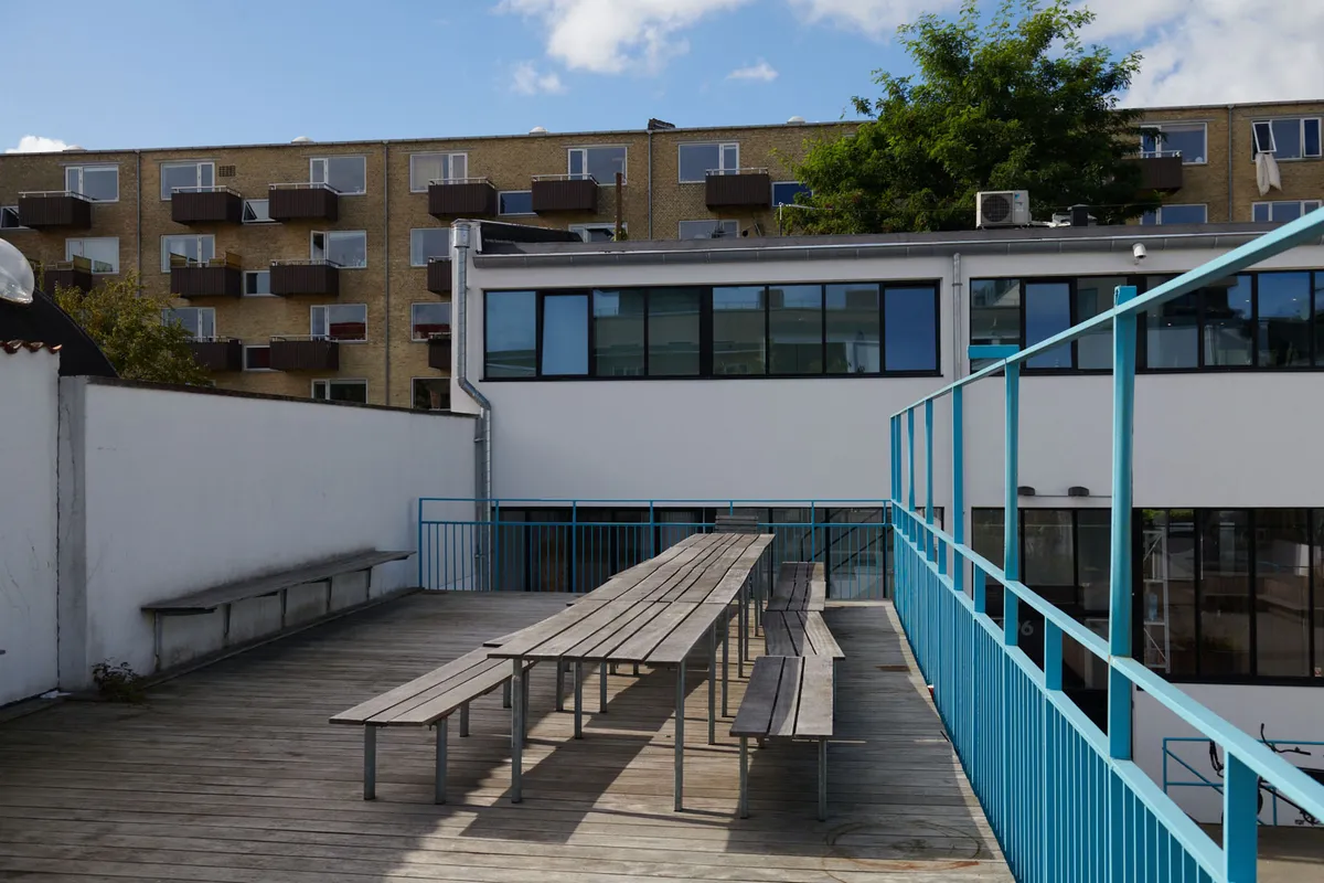 Kontorlejemål med grønne områder og flere transportmuligheder i gåafstand. Forlagshuset med lyse vinduer og terrasseområde med bænke og spisebord.