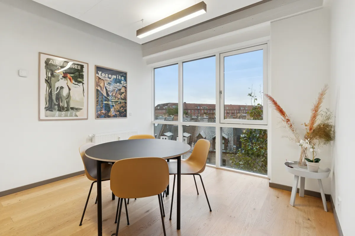 Kontorlokaler med bord, fire sæder, plante og to malerier på væggen. I baggrunden er et vindue med udsigt til en bygning. Loftet er udstyret med en lang, hvid pære.