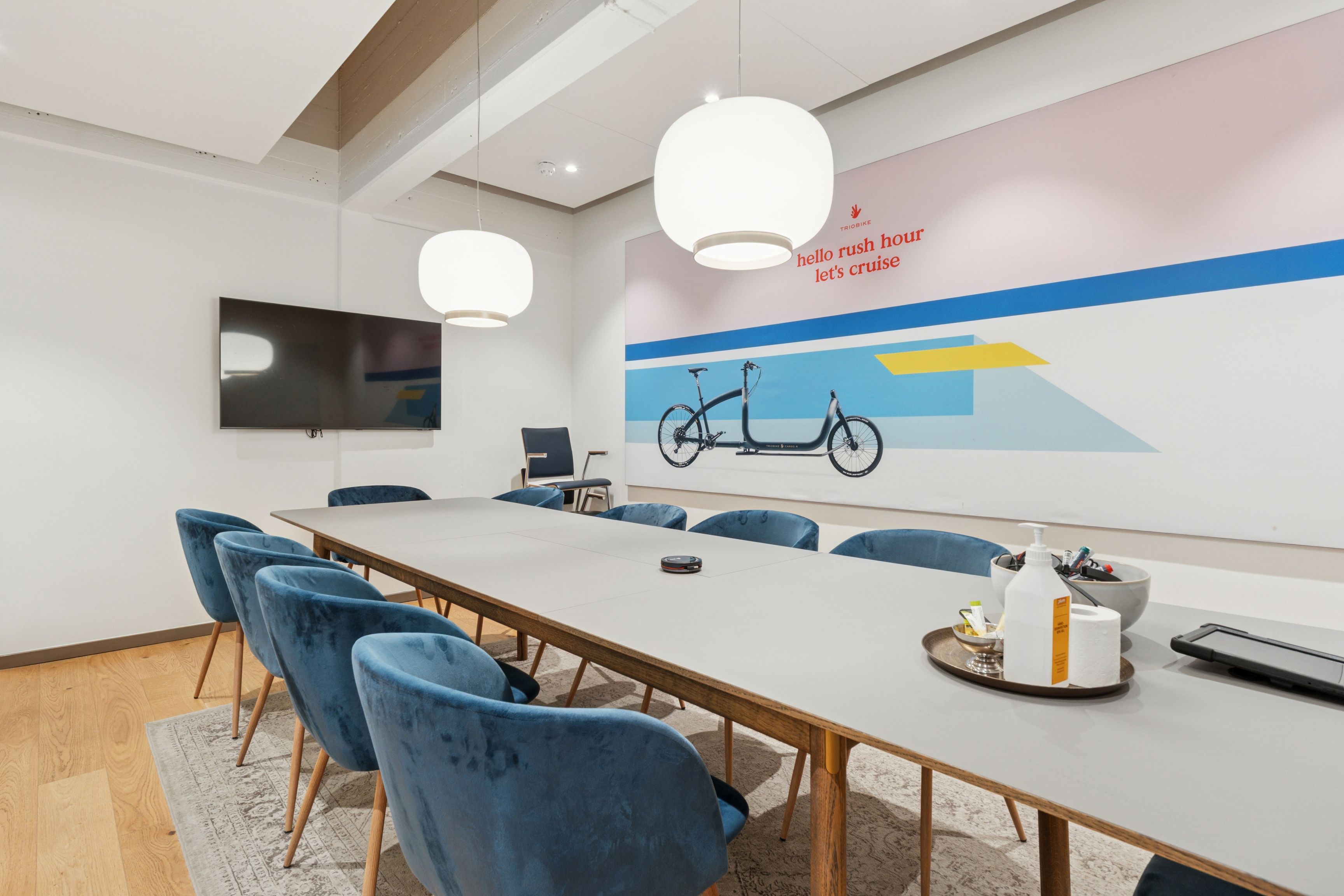 Et kontorlokale med bord, stole, tavle og tv. Loftslamper og gulvbelægning. Køkkenbord med rulle og desinfektionsmiddel.